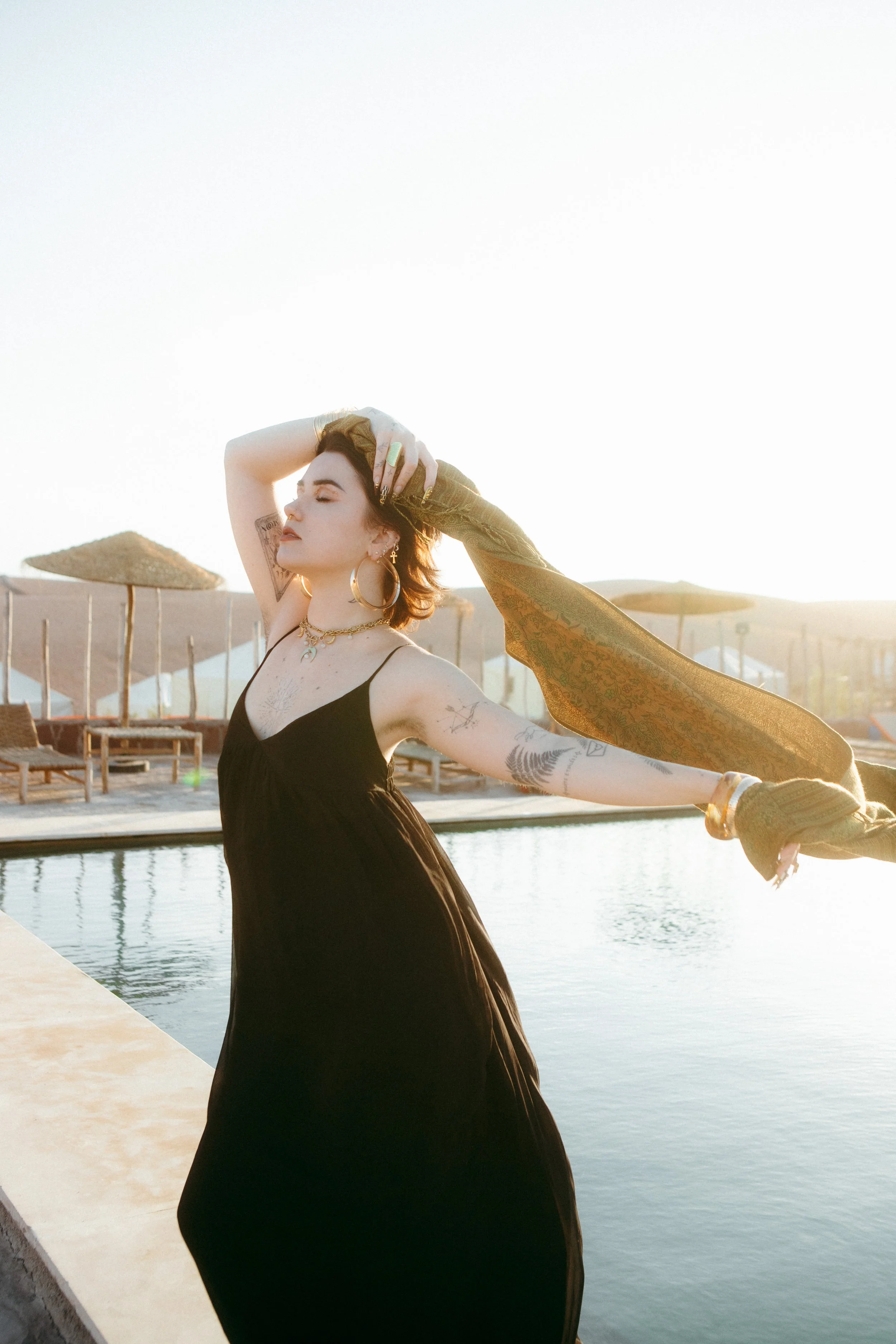 A young woman with tattoos on her arms and neck, wearing a black dress, posing outdoors near a body of water with umbrellas and chairs in the background during sunset.