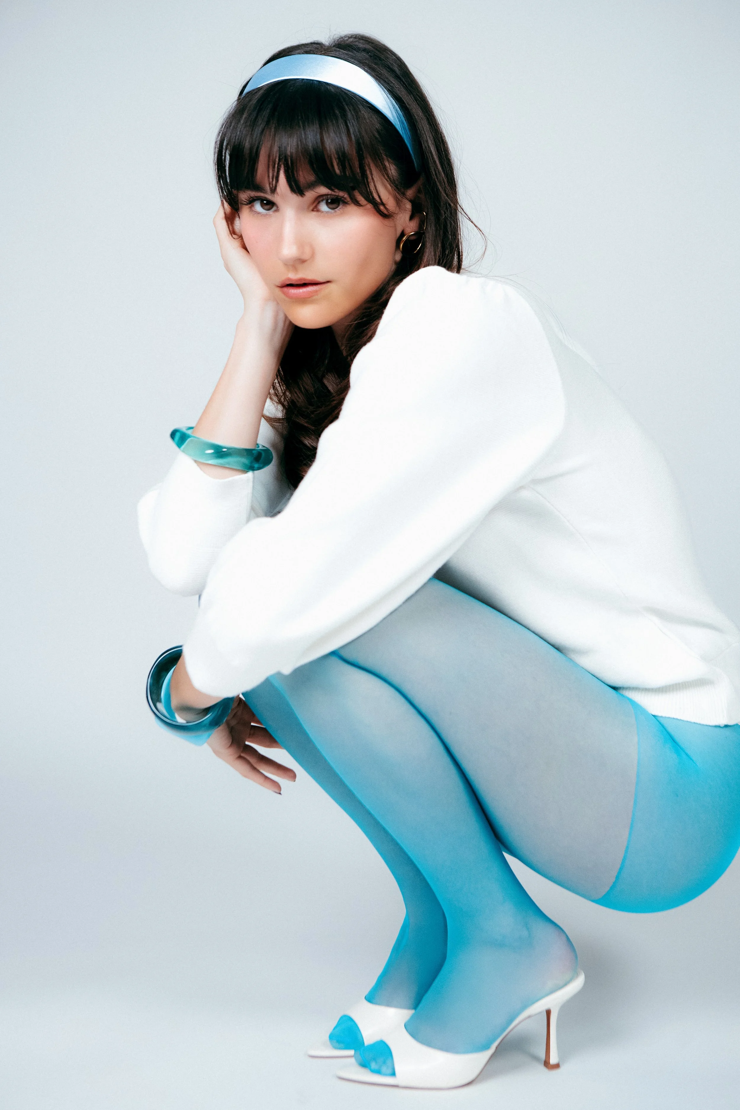 A woman with dark hair and bangs, wearing a blue headband, white top, and blue pants, sitting on a blue chair with white high heels, posing with her hand resting on her cheek.