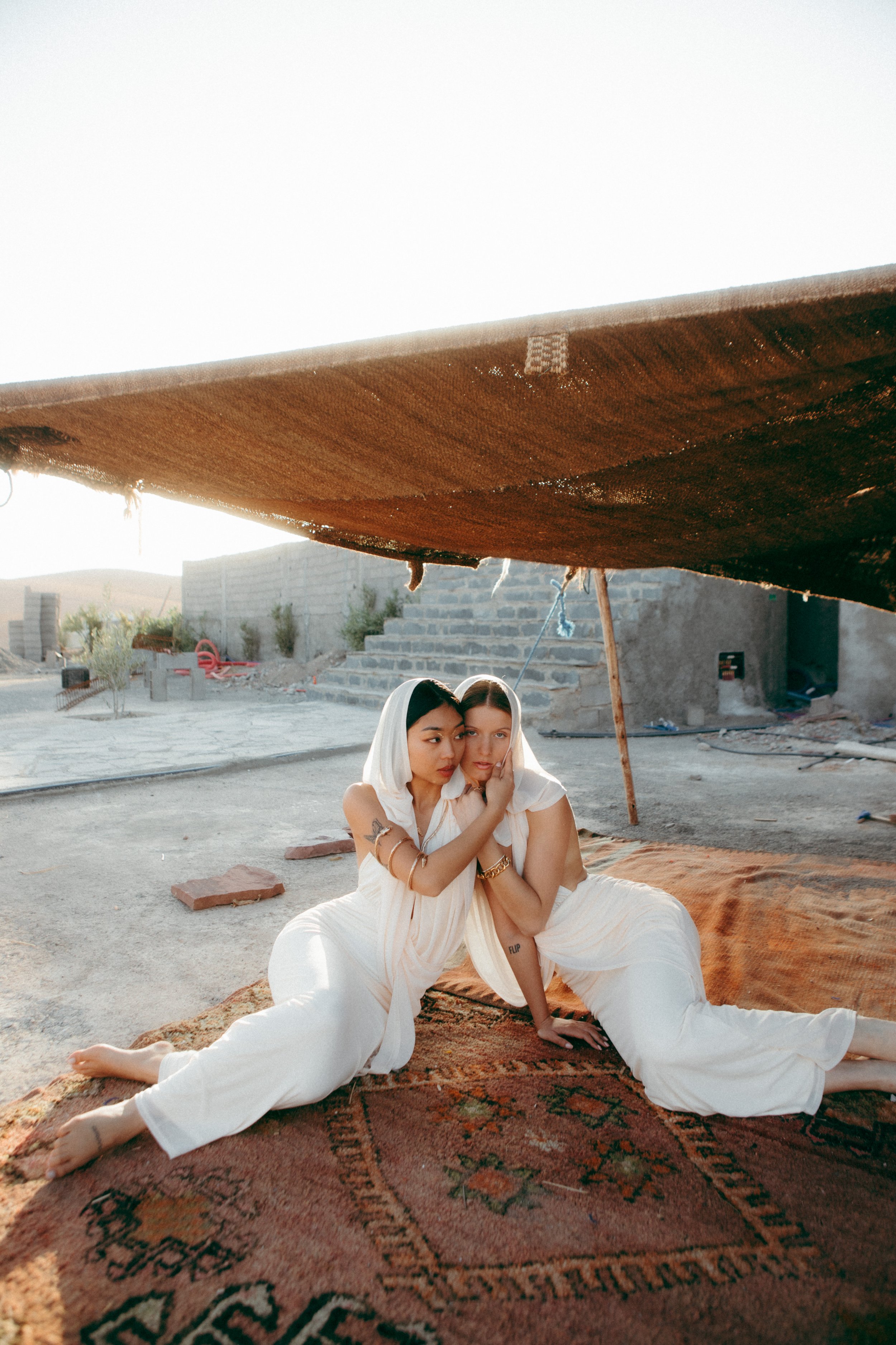 Two women sitting on a large patterned rug under a tent with a rustic outdoor backdrop, embracing each other gently and looking at each other.