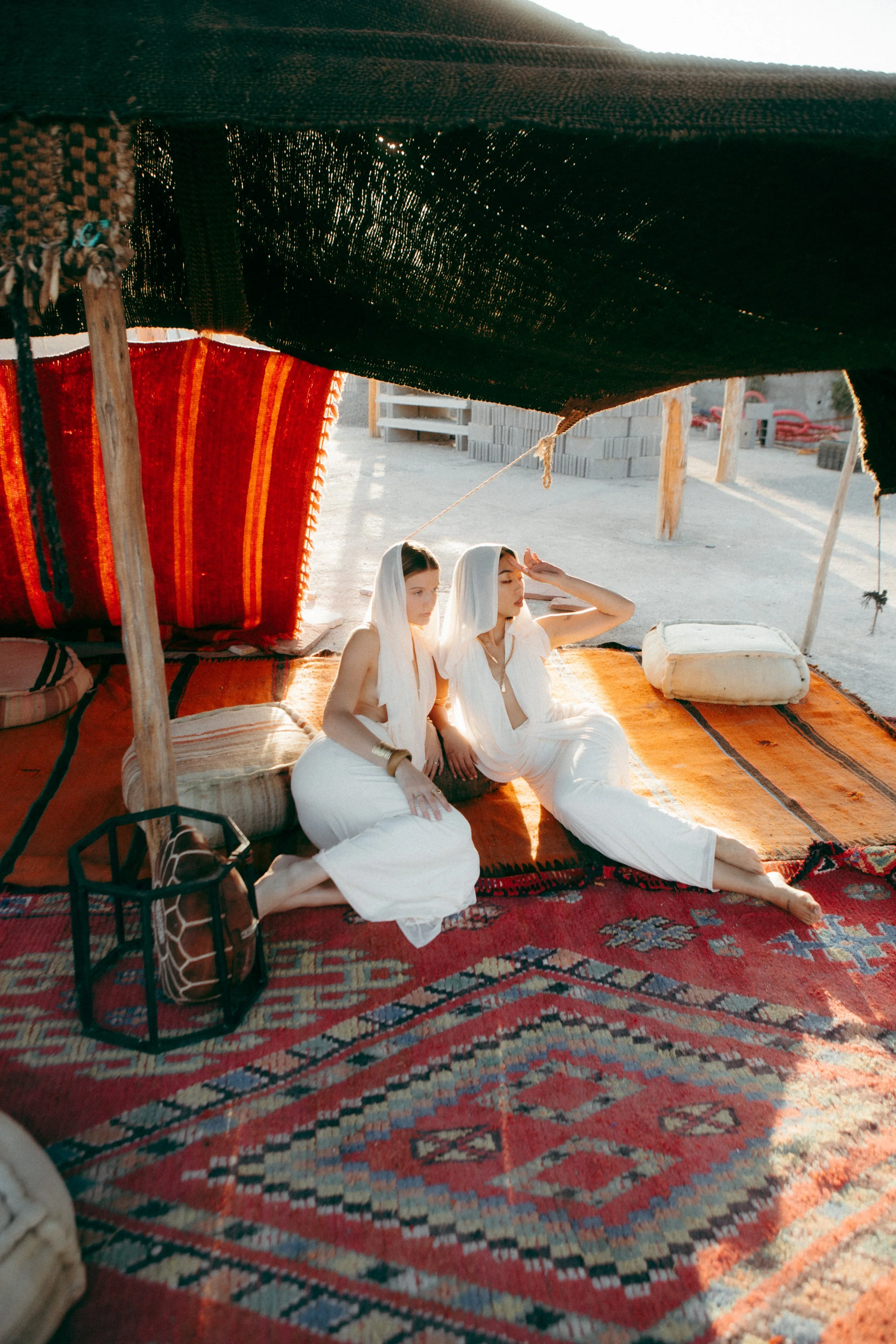 Two women sitting on patterned rugs under a tent in a desert landscape, with sandy ground, wooden supports, and construction materials in the background at sunset.