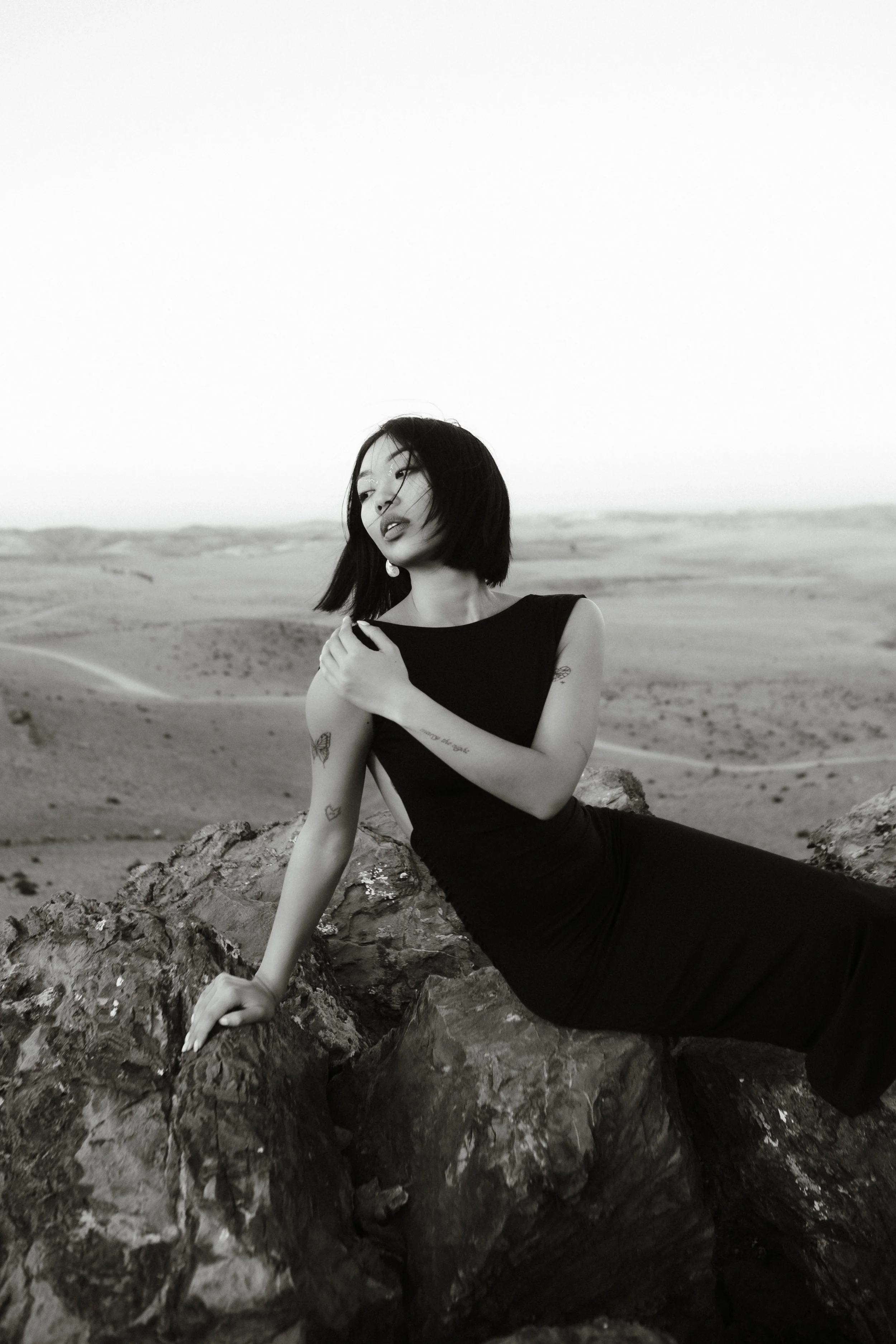 A woman with black hair in a black dress sitting on rocks in a desert landscape, looking away with wind-blown hair, in black and white photo.