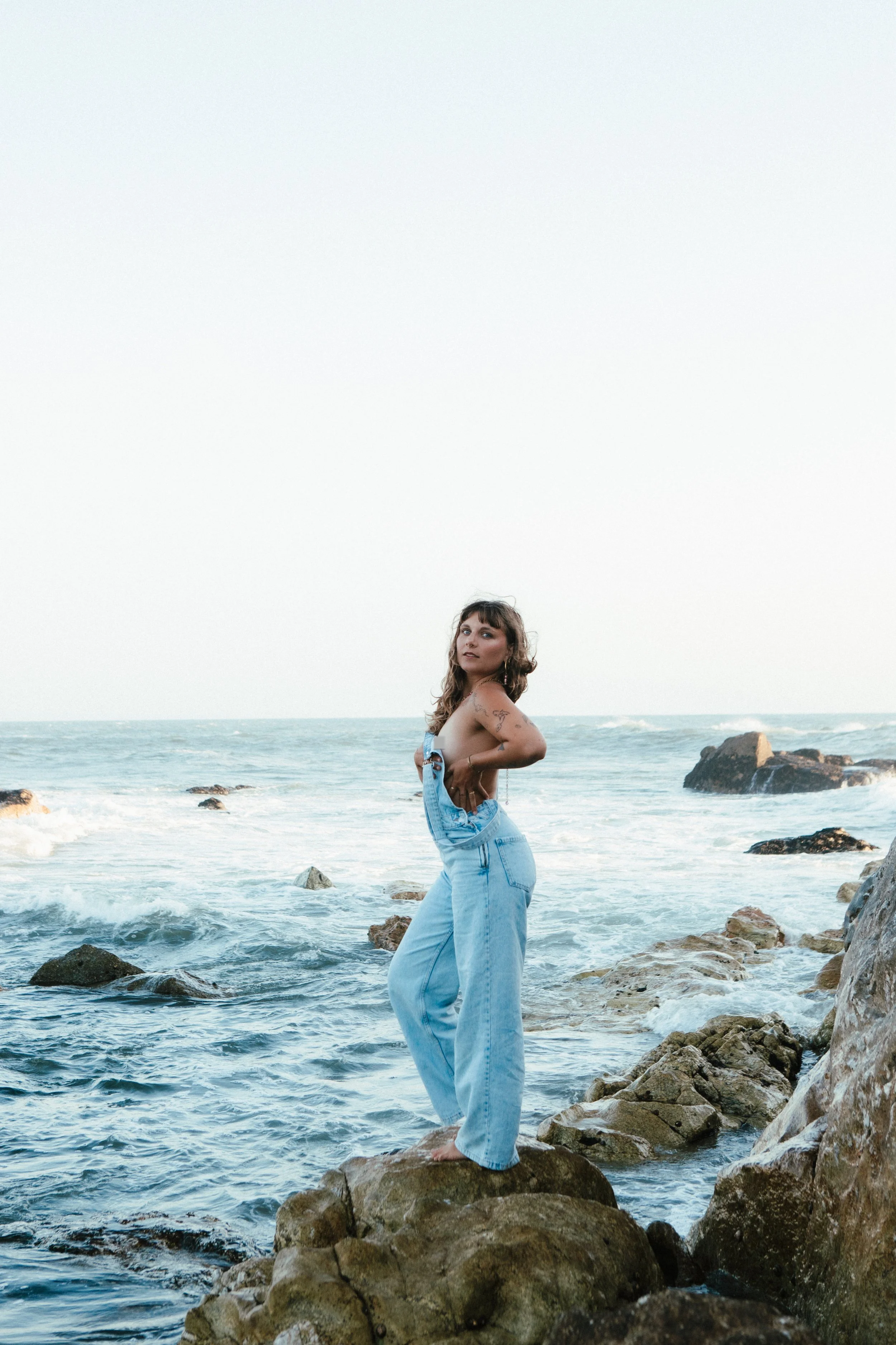 A woman standing barefoot on rocks at the edge of the ocean, wearing overalls and looking toward the camera with the sea and rocks in the background.