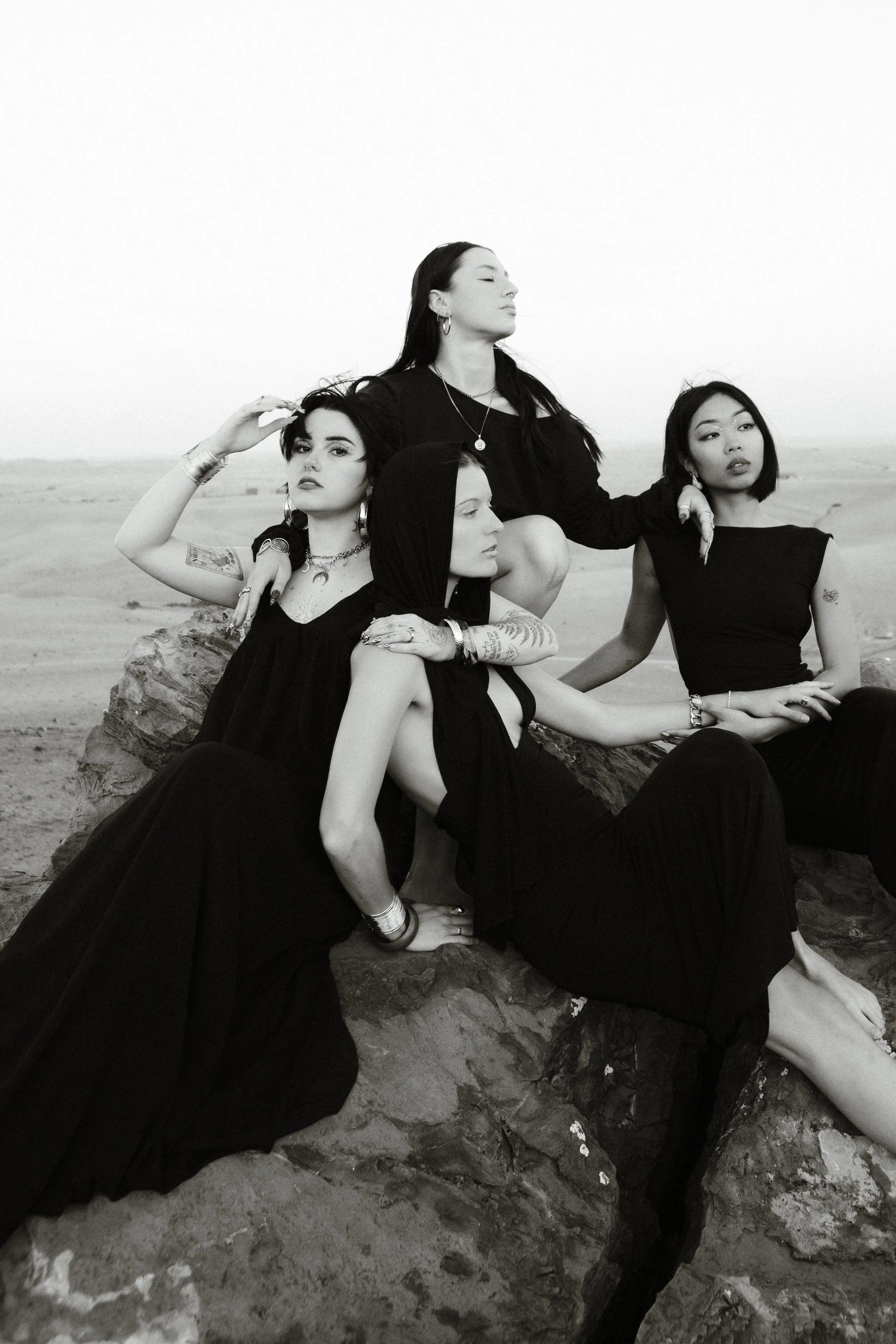 Four women sitting on rocks in a desert landscape, dressed in black clothing, looking in different directions with serious expressions, in a black-and-white photo.