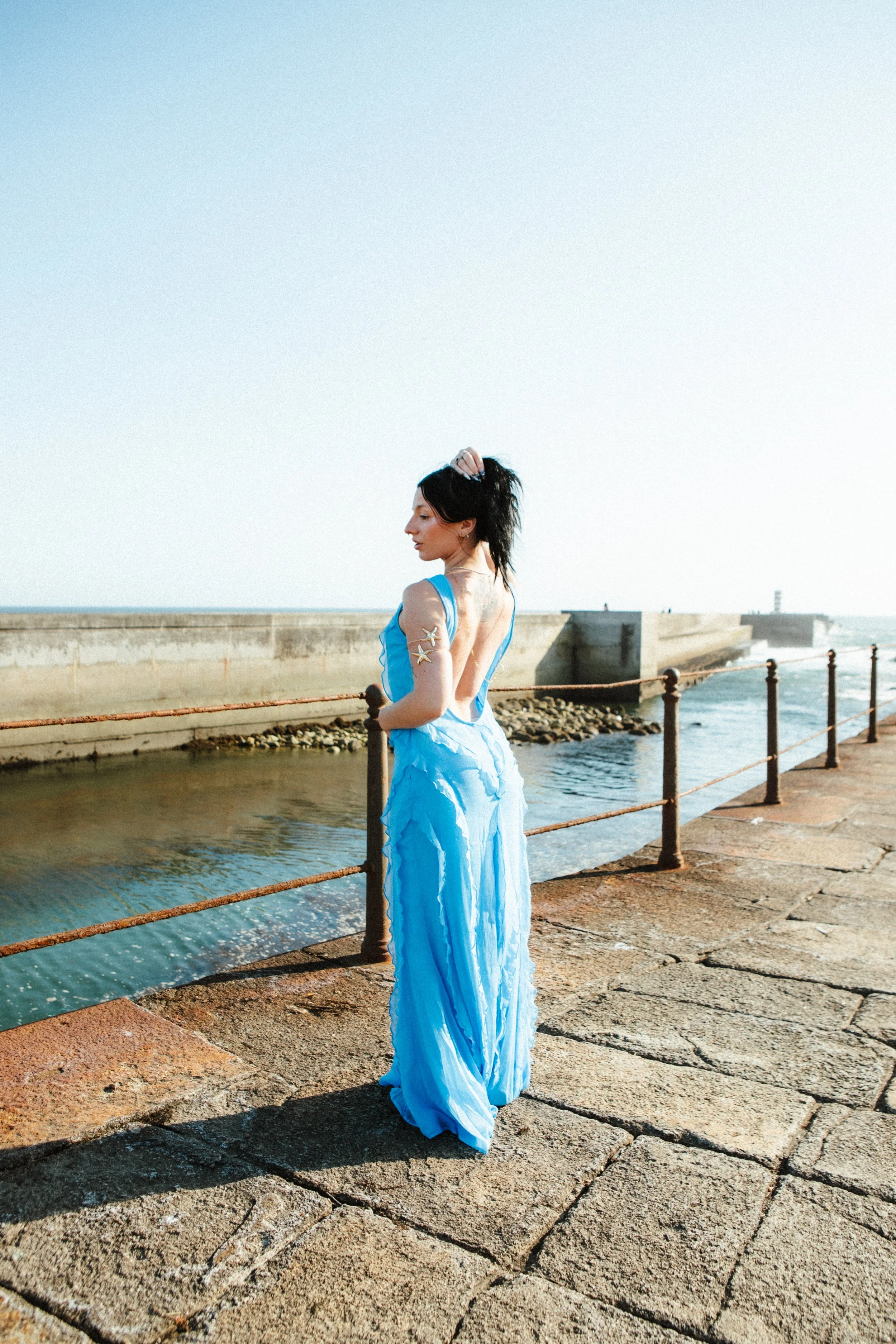 A woman with dark hair in a ponytail, wearing a flowing blue dress, stands by a waterfront on a stone walkway, with a concrete pier and railing in the background, and the ocean stretching out behind her.