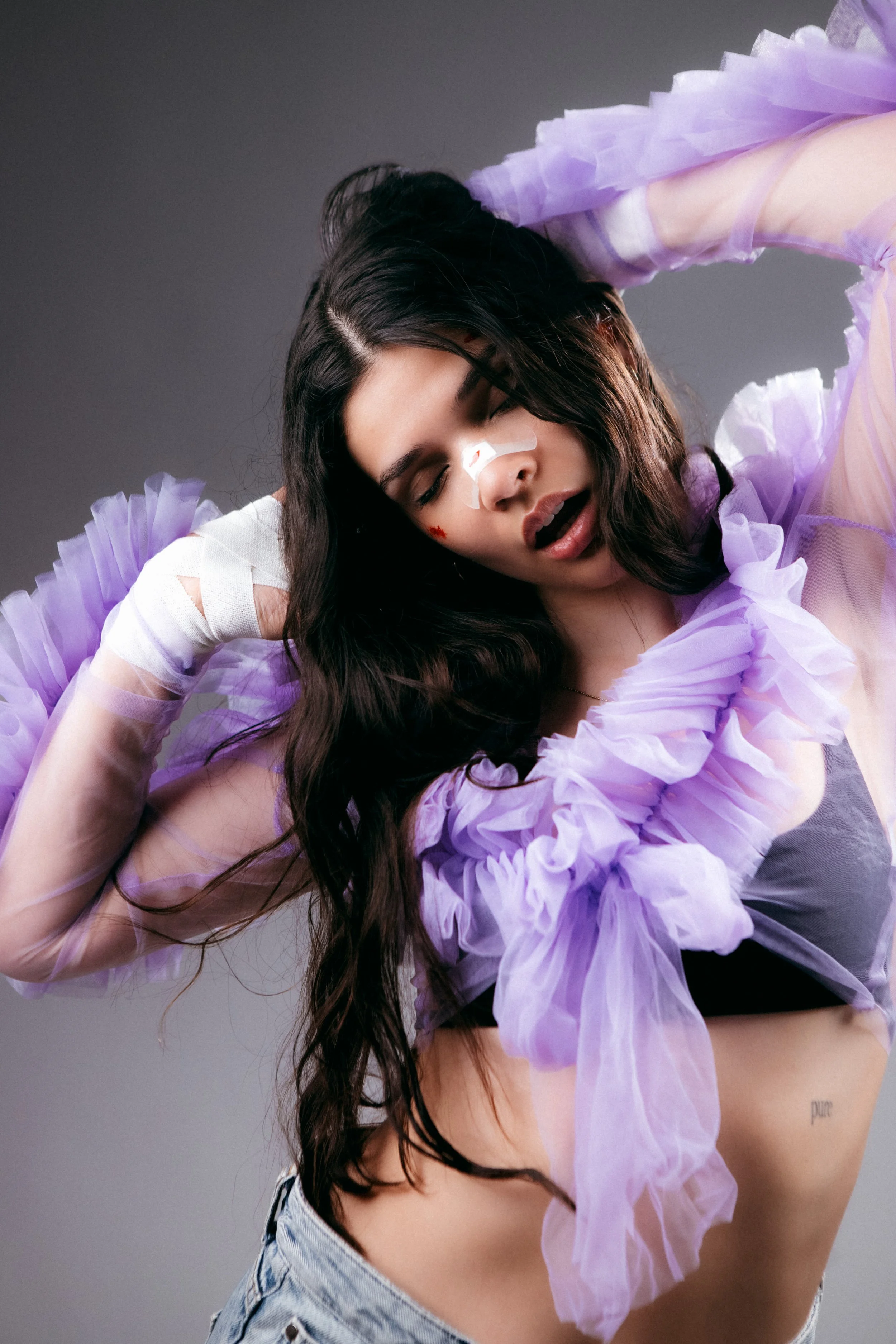 A woman with long dark hair and a nose injury, wearing a light purple ruffled top, with her eyes closed and mouth open, posing against a gray background.
