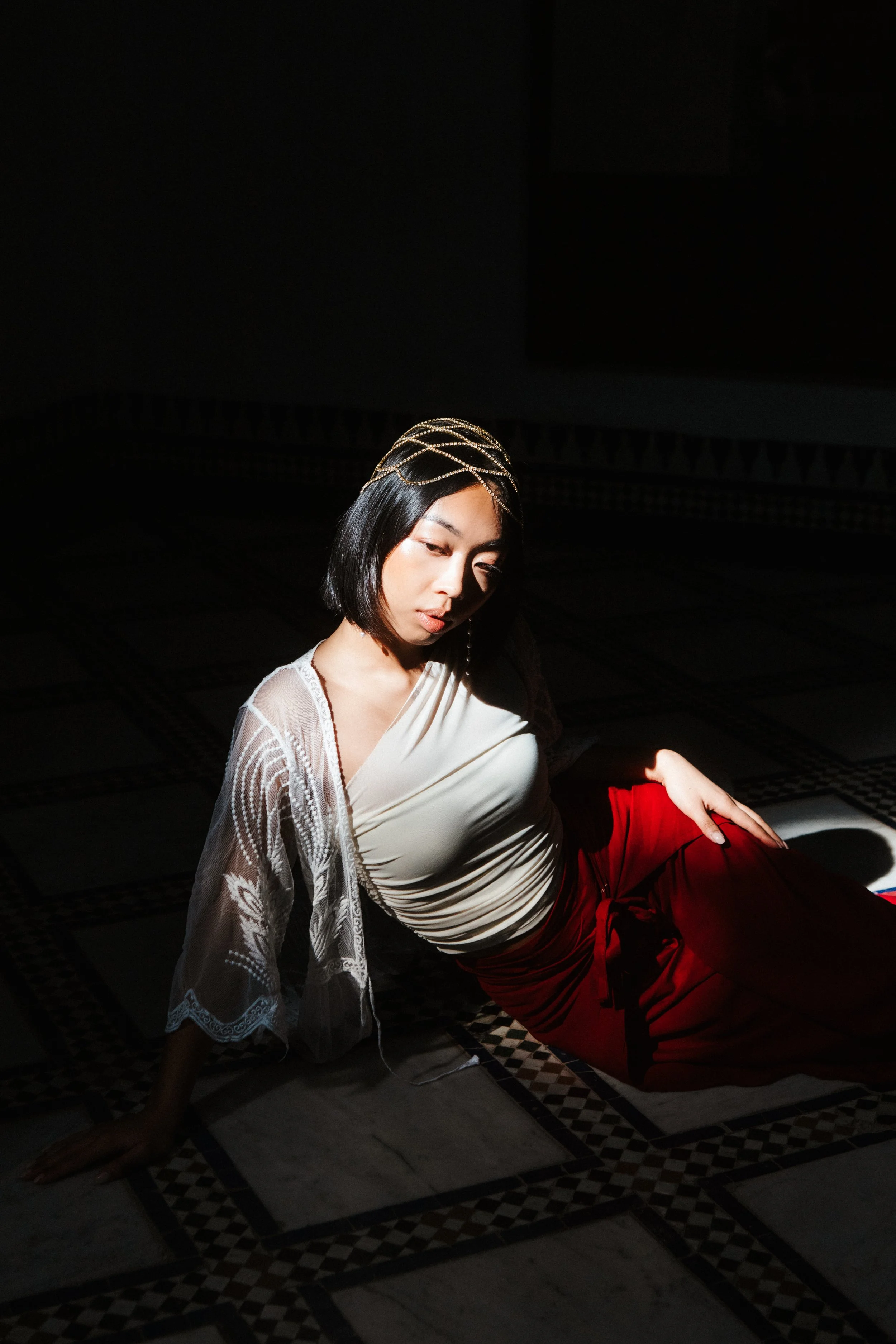 A woman with short black hair, wearing a white draped top and a sheer lace shawl, is sitting on the floor with one arm extended behind her. She has a headpiece with gold accents and is partially illuminated by a light source, creating a dramatic, sha