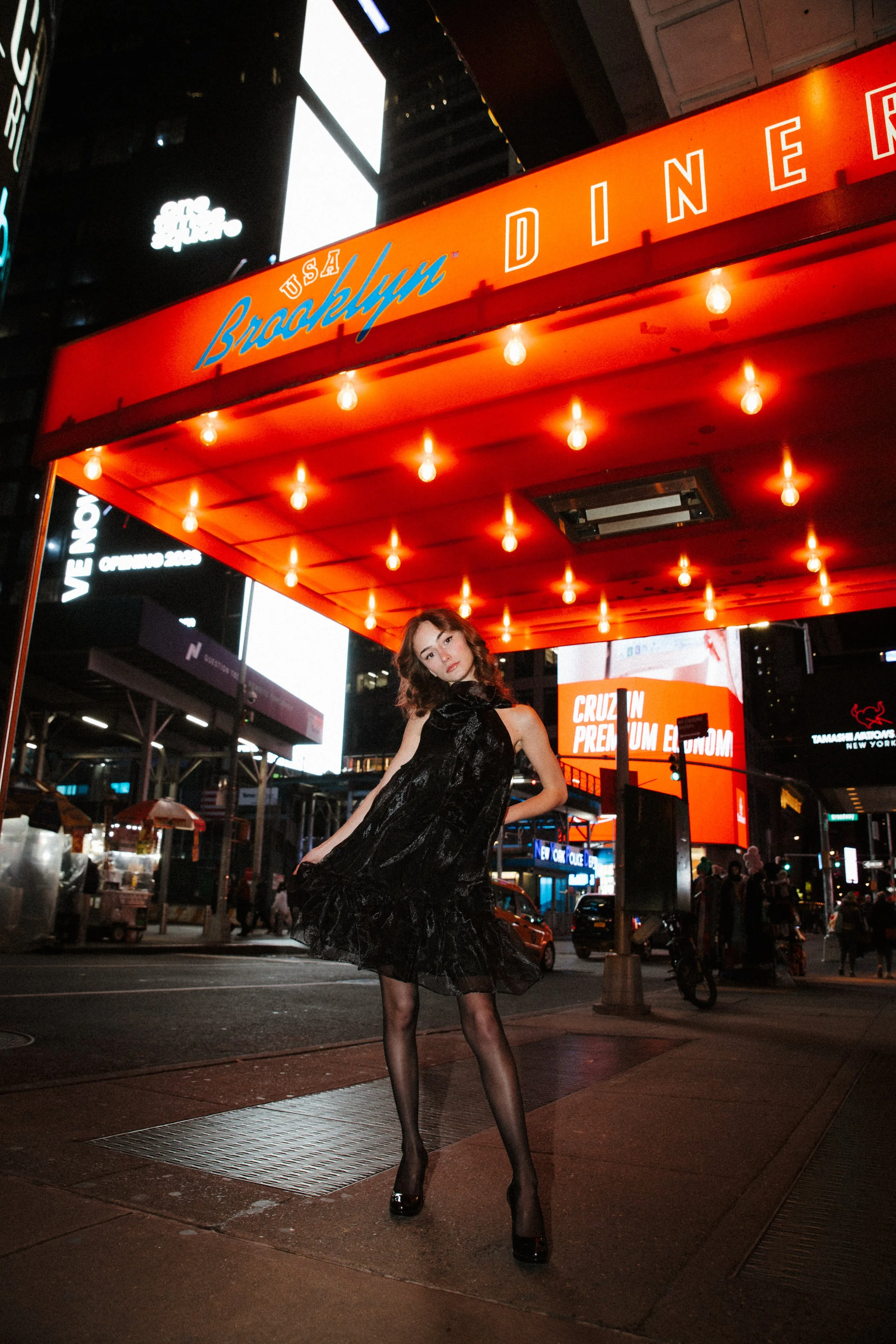A woman in a black dress with a tulle skirt posing on a city street at night, illuminated by bright signs and lights. Neon signs and billboards are visible in the background.