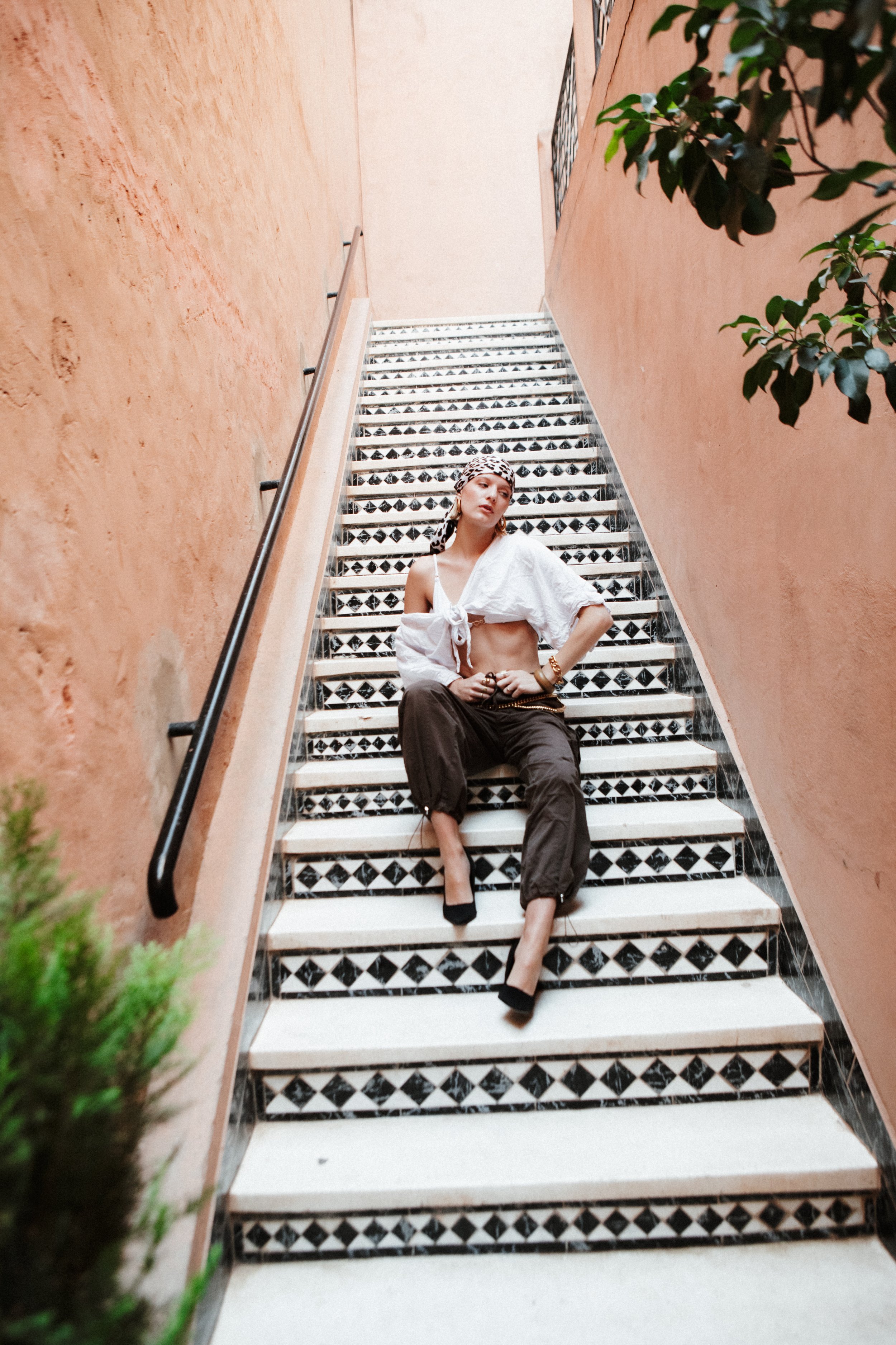 A woman sitting on patterned black and white tiled stairs with beige walls on either side, wearing a white top, brown pants, black high heels, a leopard print headscarf, and jewelry, with greenery on the left side.