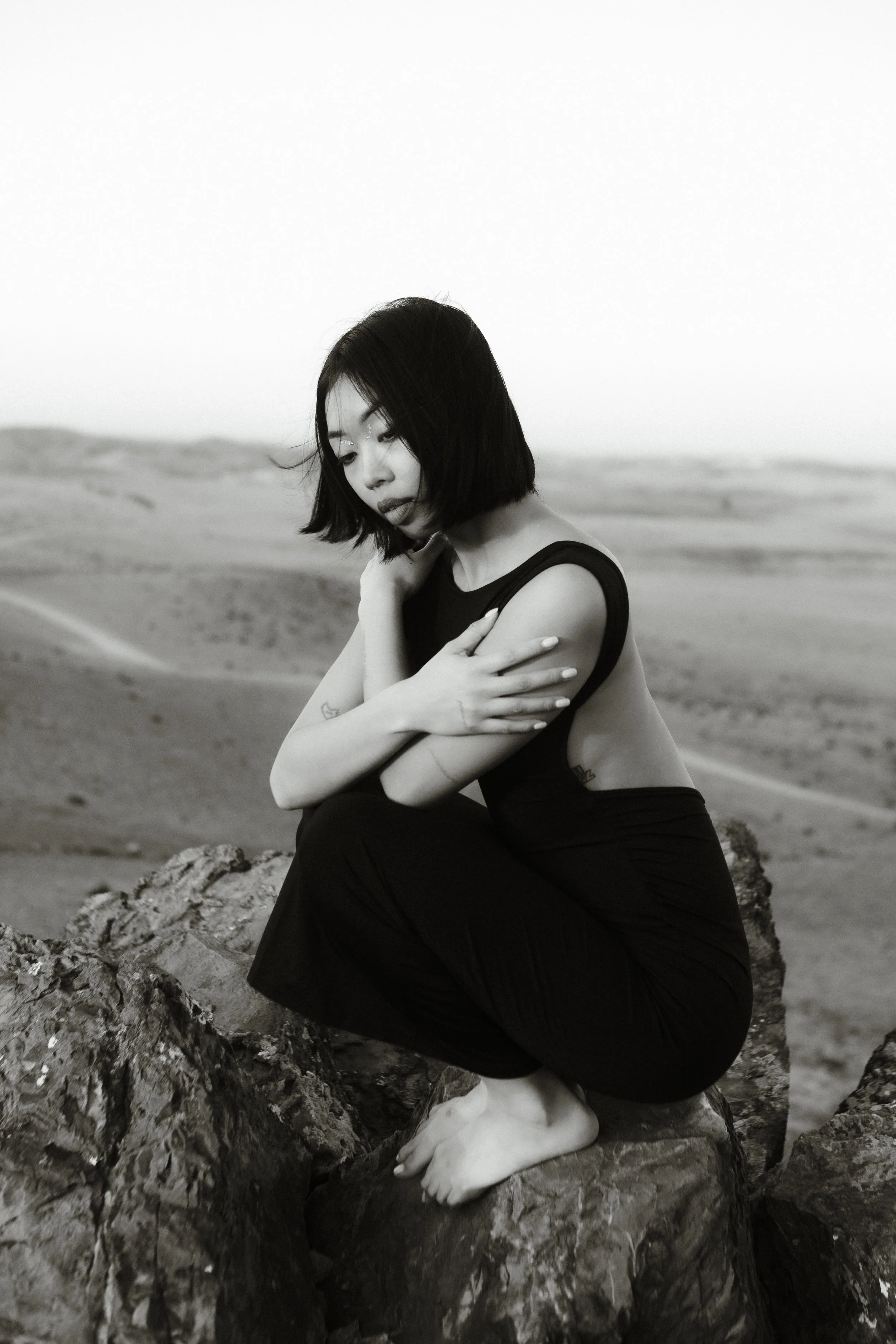 A woman with short black hair, wearing a sleeveless top and dark pants, sitting on rocks in a desert landscape, with her eyes closed and arms crossed over her chest.