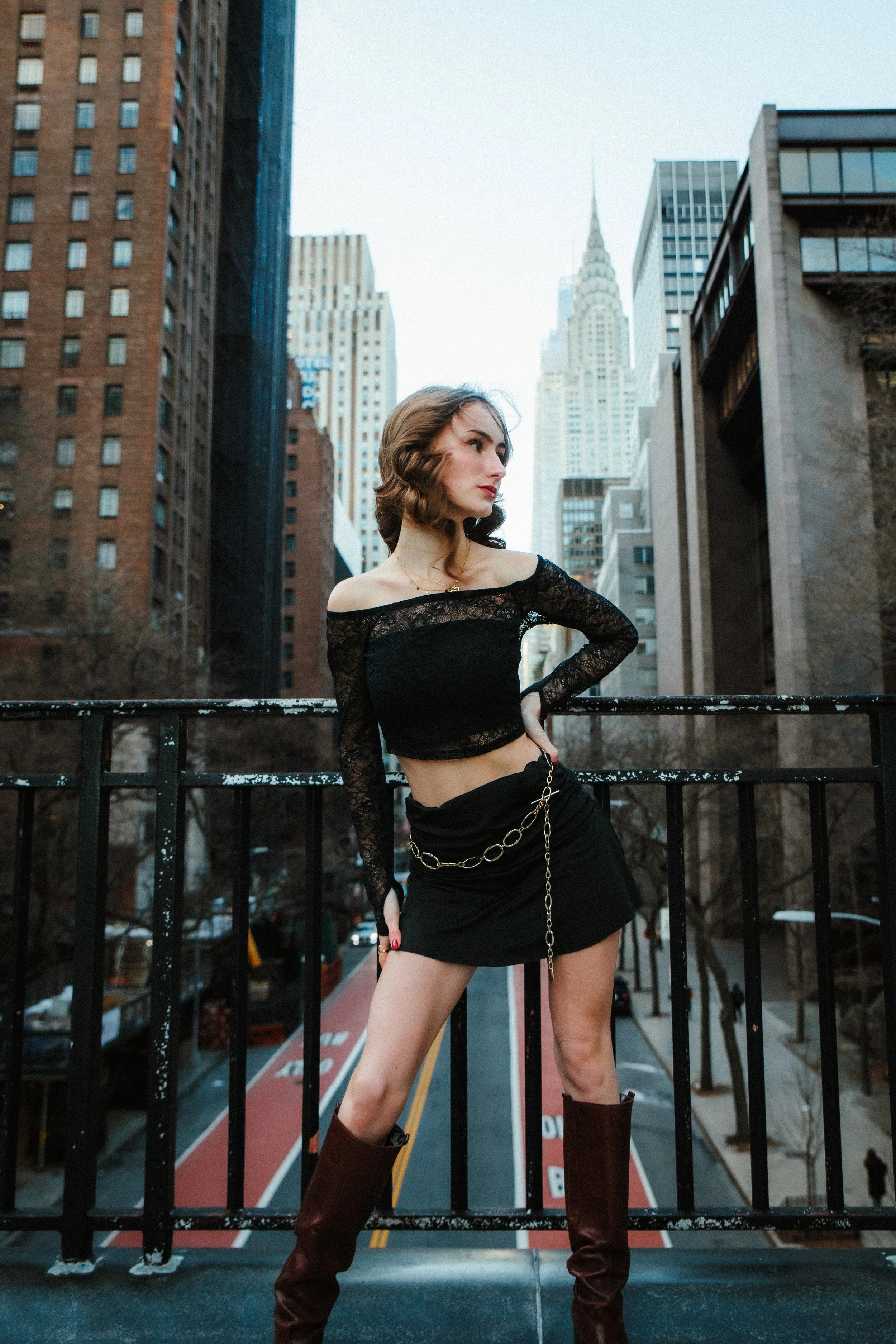 A young woman with shoulder-length hair wearing a black lace off-shoulder top, black skirt with a chain belt, and knee-high maroon boots, posing on a city street with tall buildings and a famous skyscraper in the background.