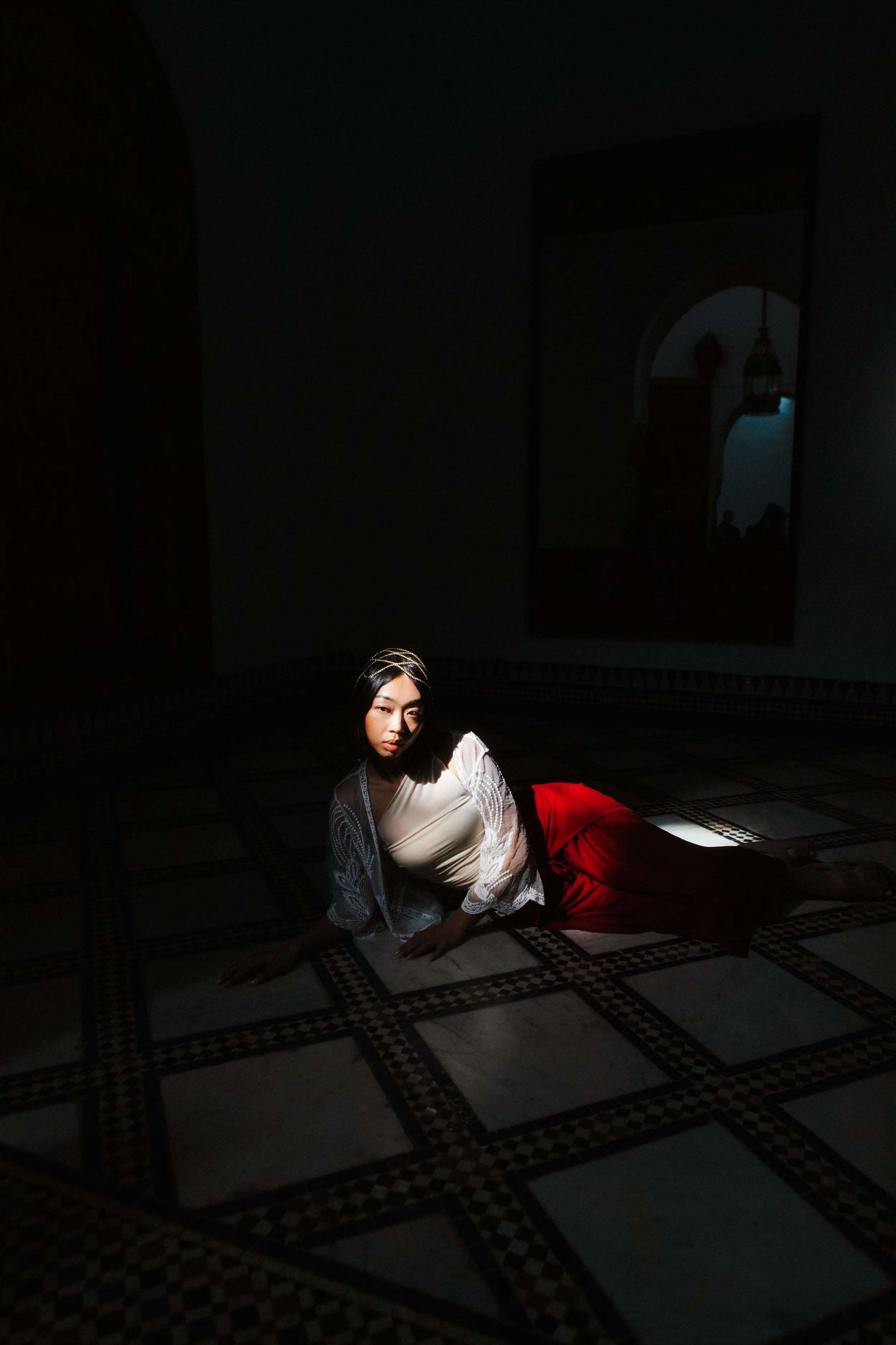 A woman dressed in elegant clothing, with a white lace top and red skirt, lying on the patterned floor of a dimly lit room with a mirror and lantern in the background.