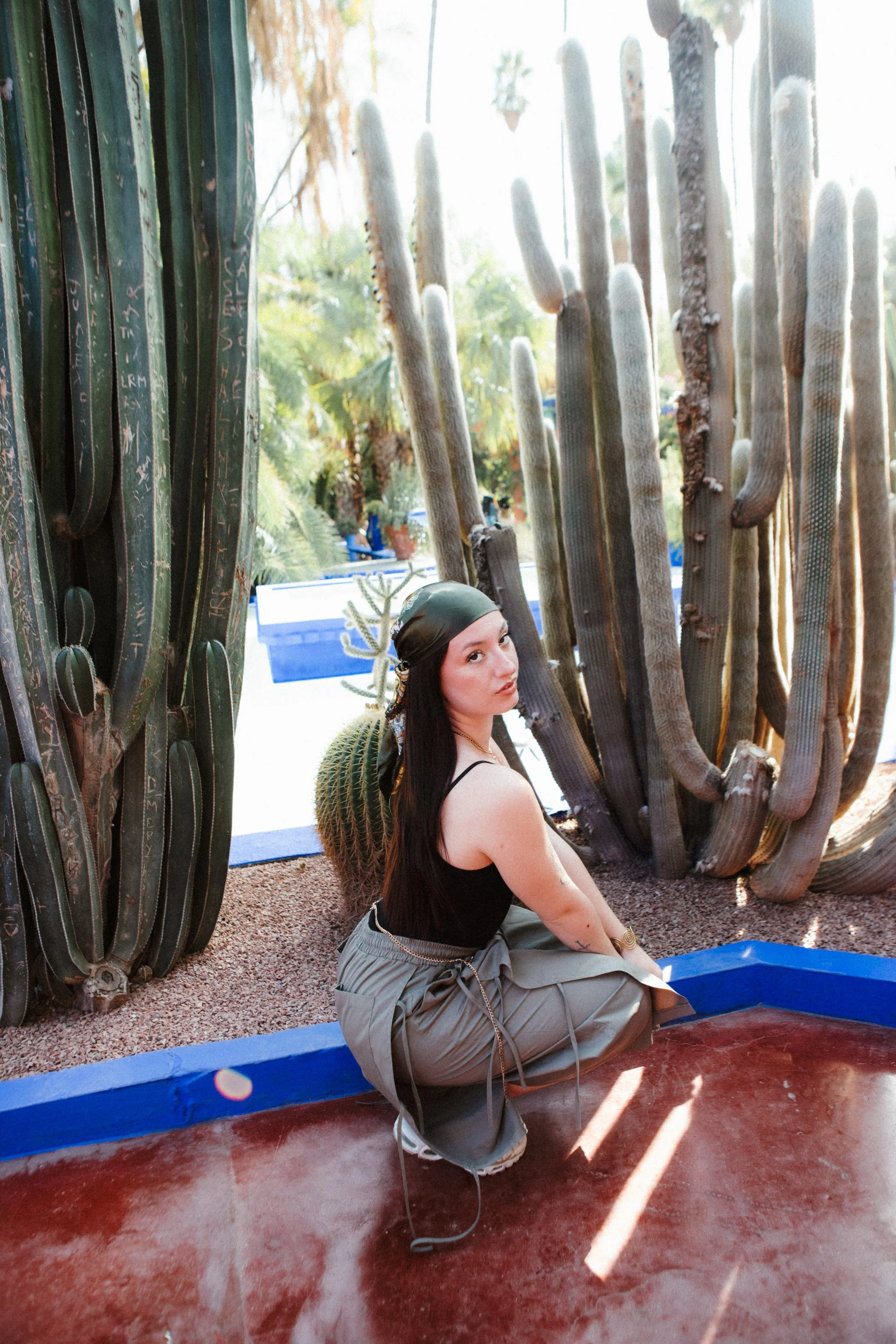 A woman with long dark hair, wearing a black tank top, loose gray pants, a green headscarf, and white sneakers, kneeling on reddish ground in front of tall, cactus plants outdoors during daytime.