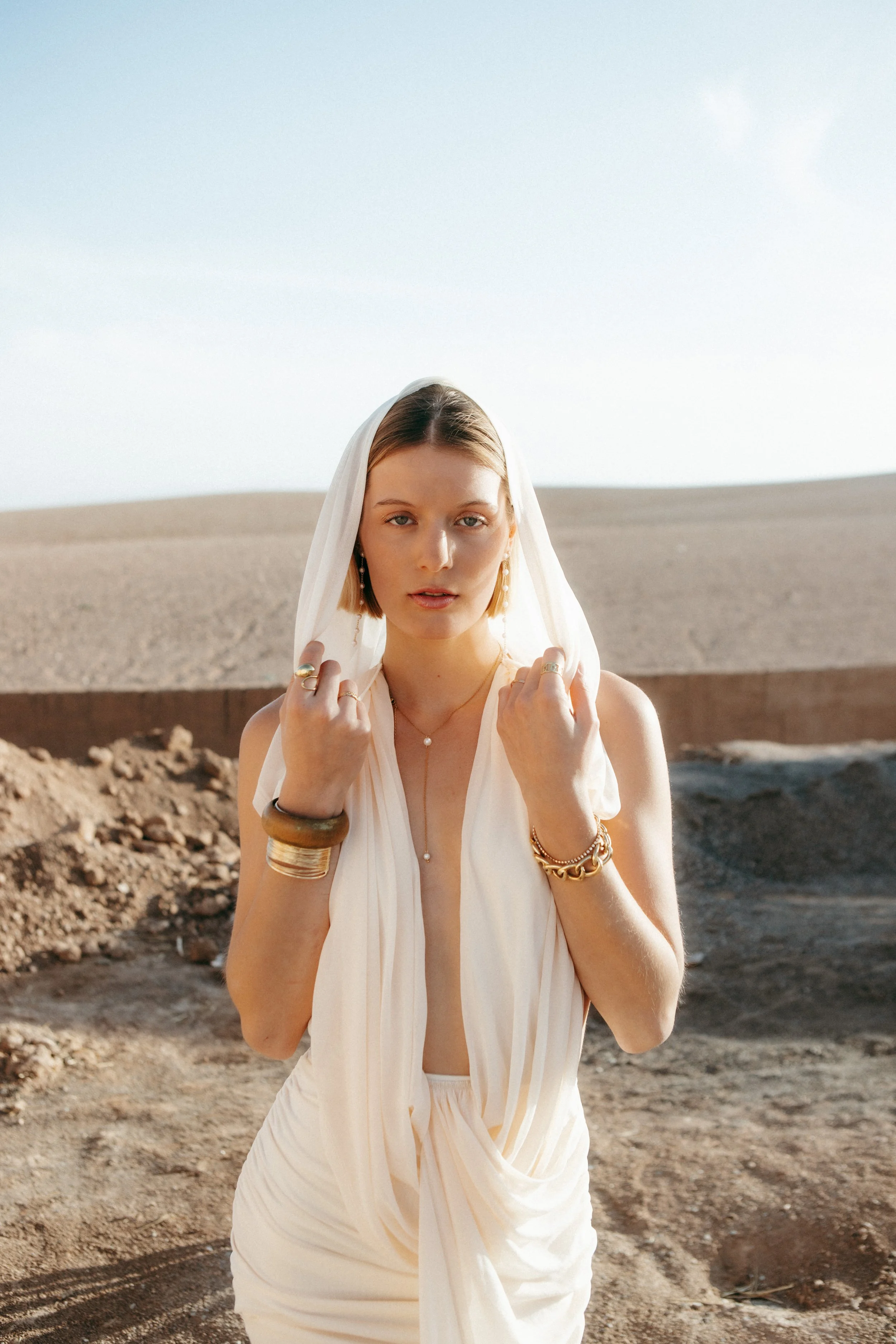 A woman standing outdoors in a desert-like landscape, wearing a white dress with a plunging neckline and a sheer white veil. She has short blonde hair, earrings, and jewelry on her hands and wrists, and is holding the veil with both hands. The sky is
