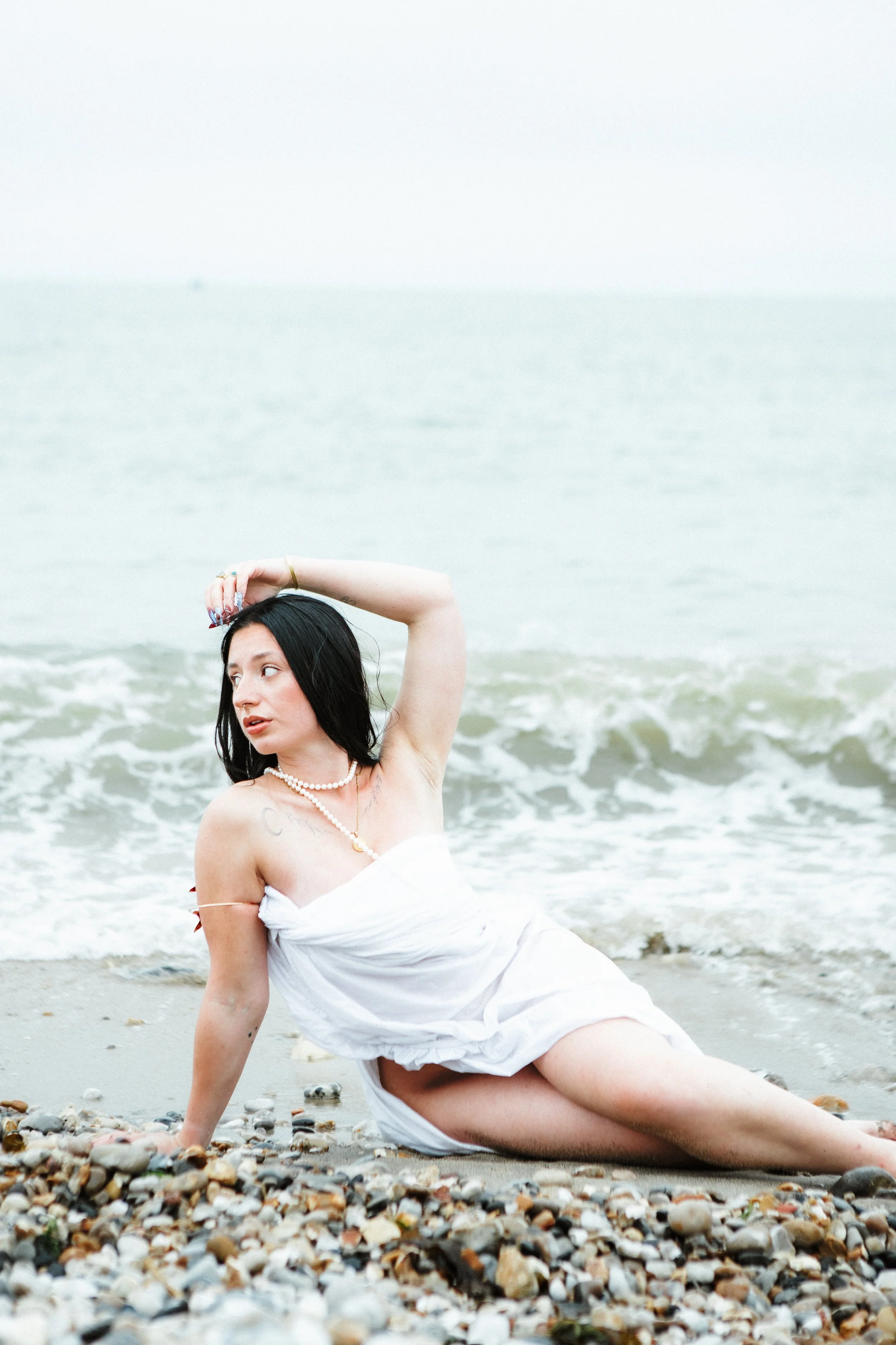 A woman with black hair at the beach, wearing a white strapless dress and jewelry, sitting on the pebbled shore near the ocean, with her arm raised and looking to the side.