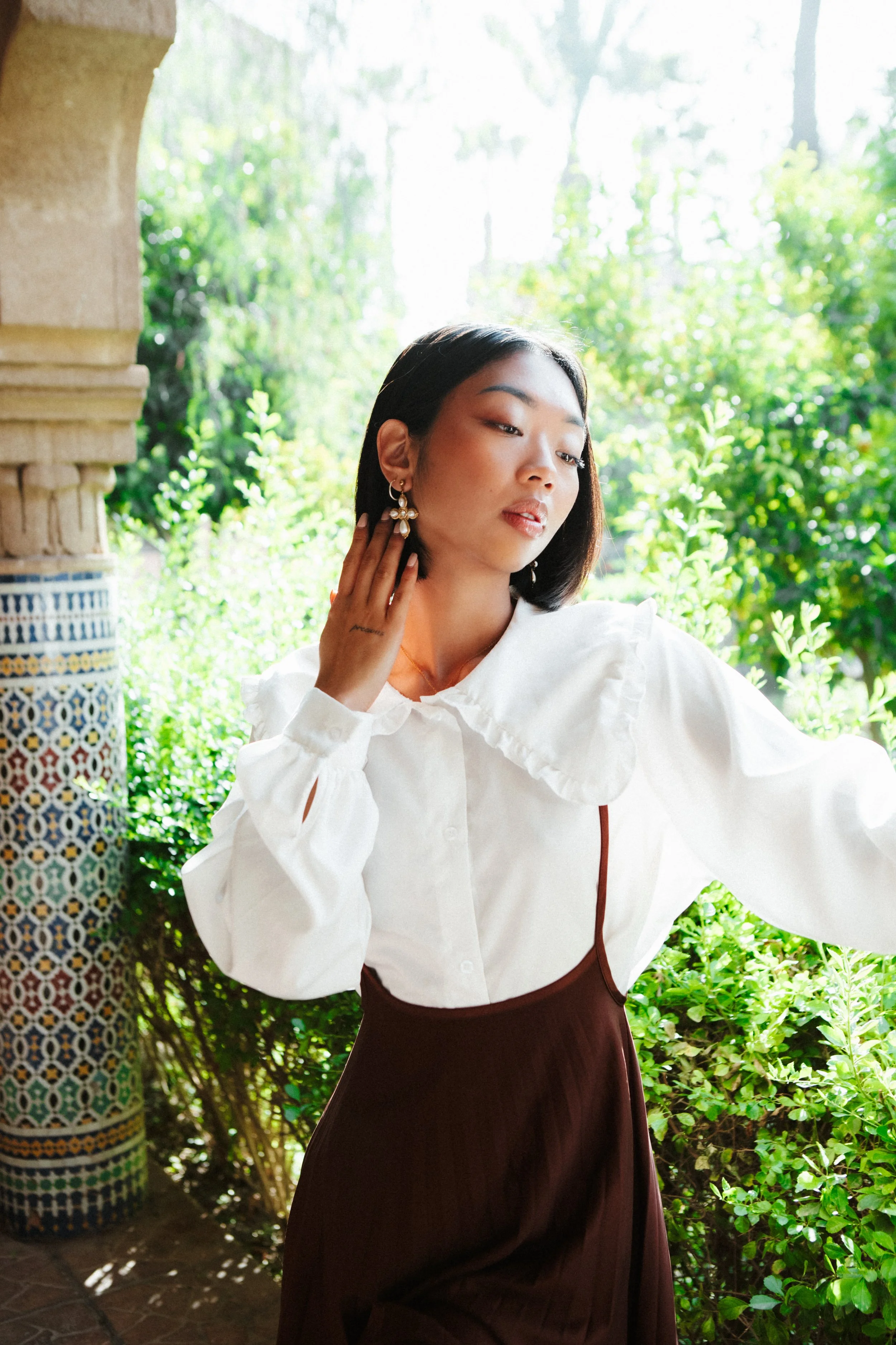 A woman with black hair wearing a white blouse and brown skirt standing outdoors next to a decorated column, touching her ear, with lush green plants and sunlight in the background.