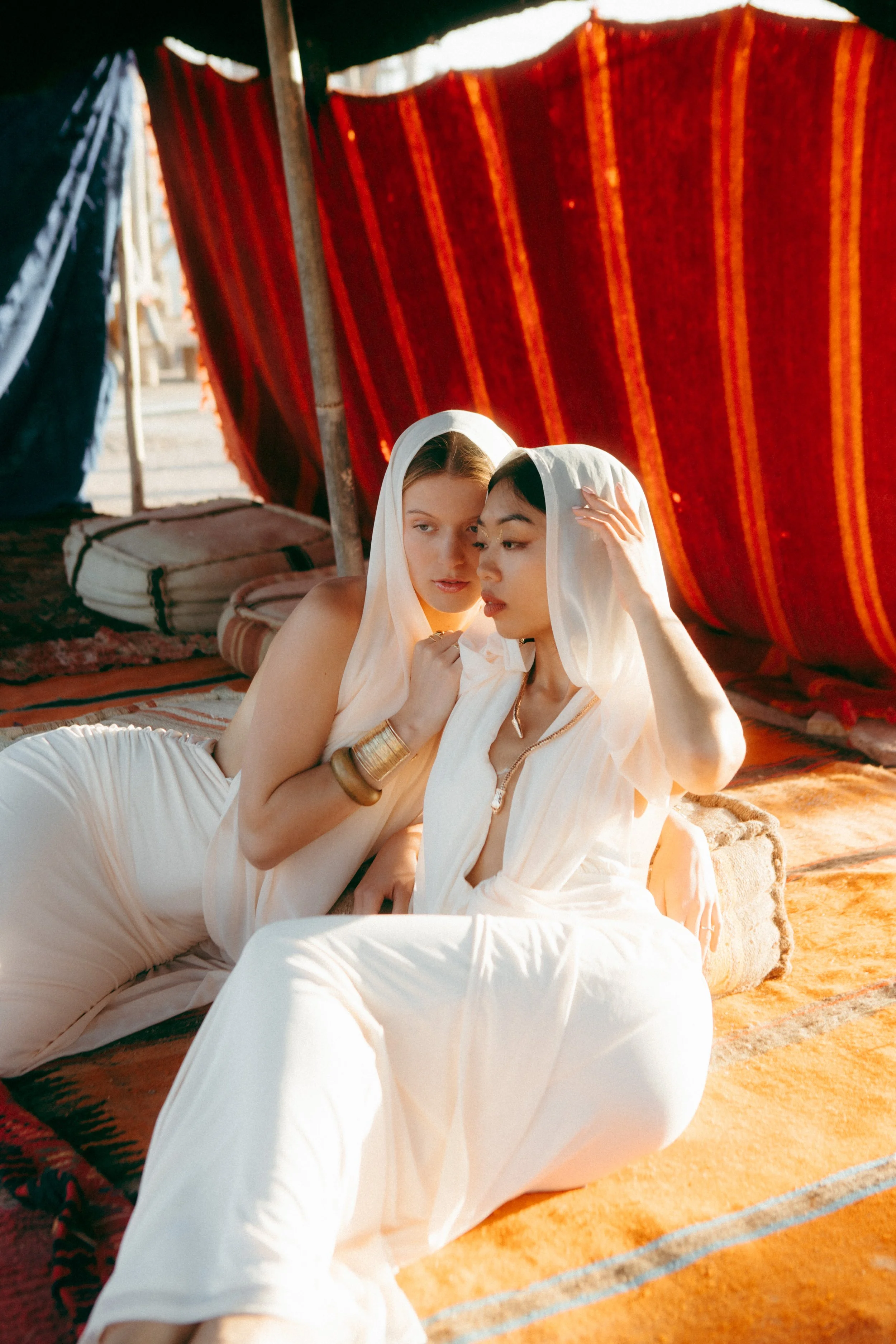 Two women dressed in white robes sitting on a colorful rug under a tent made from red and blue fabric, with one adjusting the other's headscarf, in a desert setting.