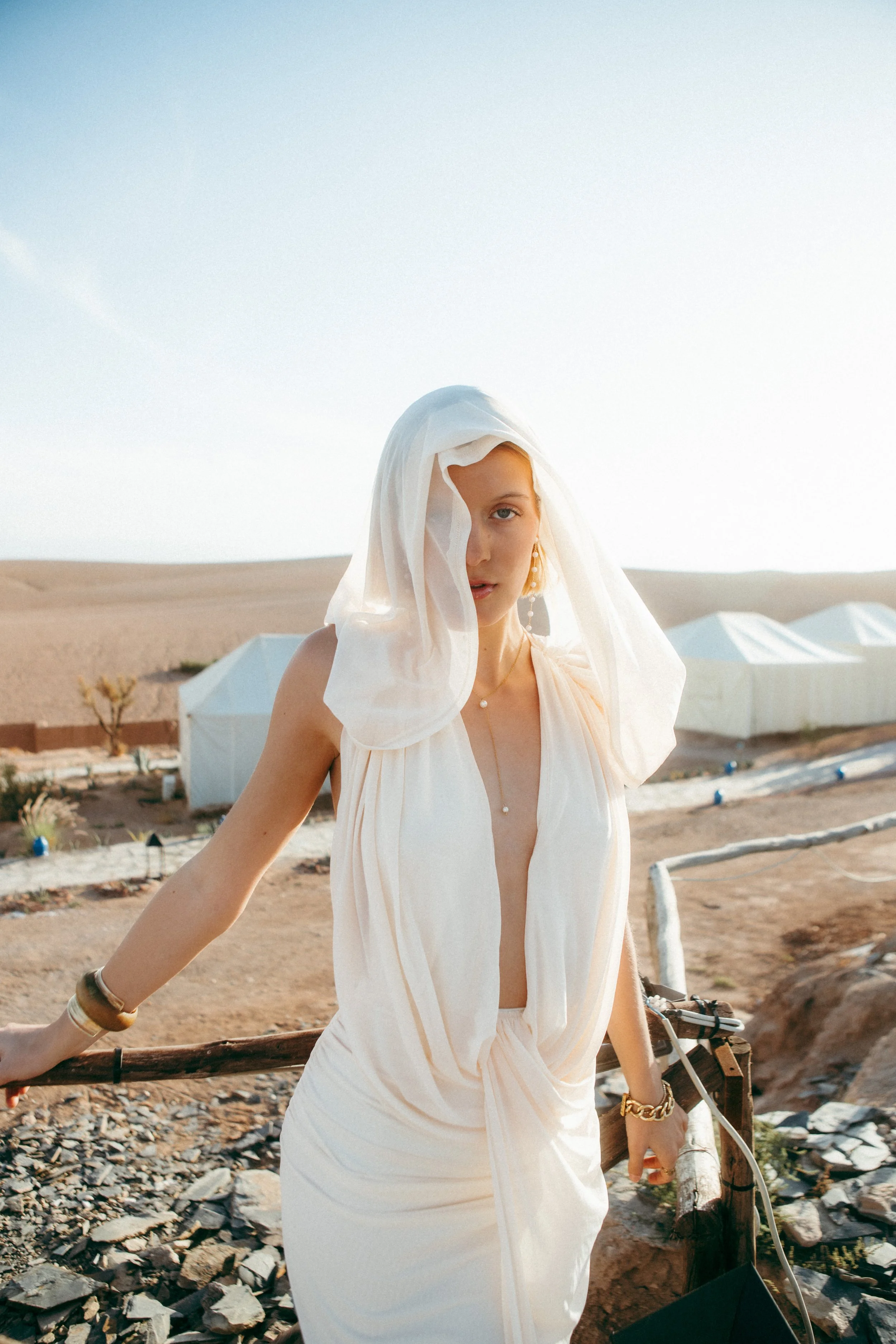 A woman dressed in a flowing white outfit with a sheer white veil over her head, standing outdoors in a desert-like environment with tents in the background.