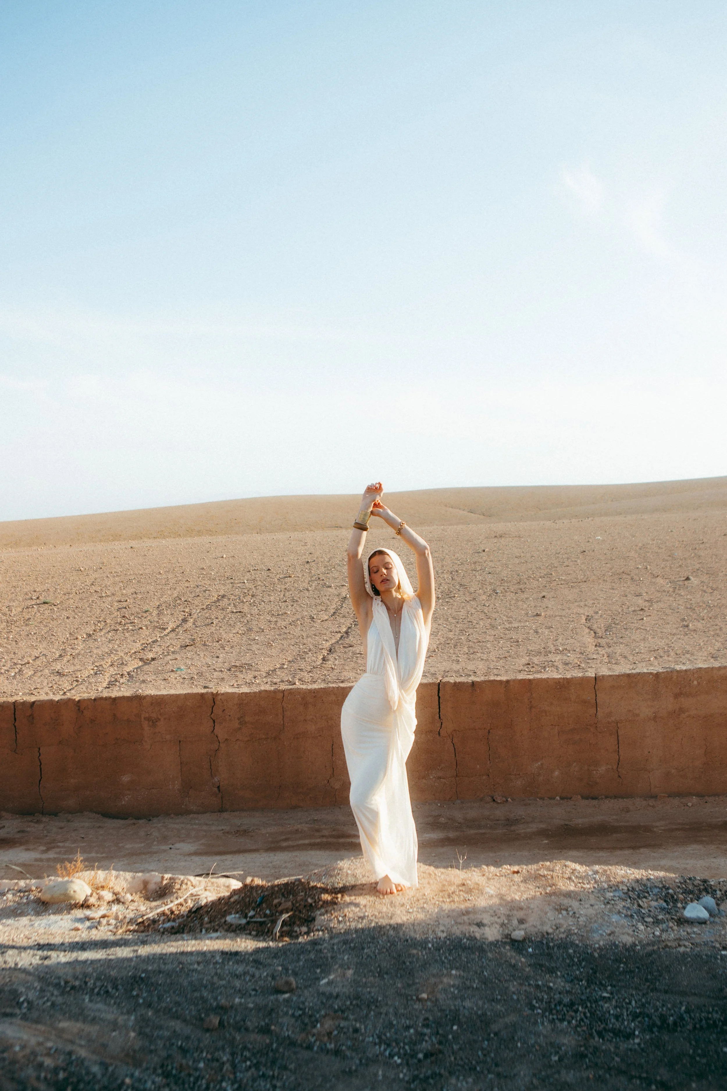 A woman in a flowing white dress stands barefoot on a concrete ledge in a desert landscape, with her arms raised above her head, holding a bracelet. The background includes a clear sky and sandy terrain.