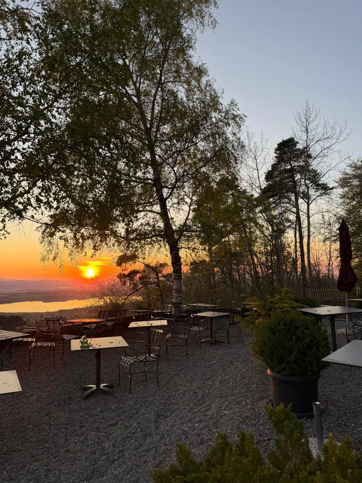 Sonnenuntergang über einem Teich, im Vordergrund sind Tische und Stühle im Freien, umgeben von Bäumen.