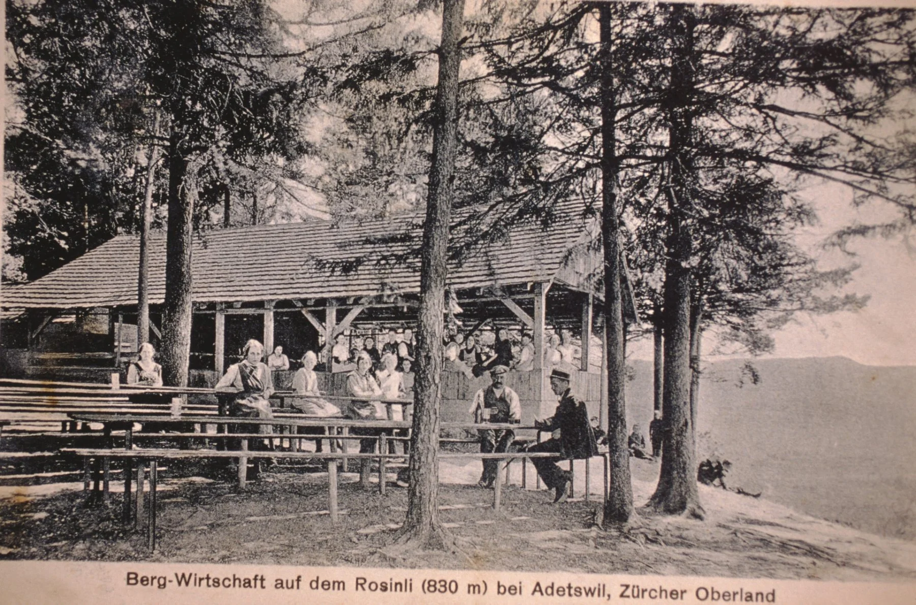 Historisches Foto einer Bergwirtschaft auf dem Rosinli bei Adetswil, Zürcher Oberland, umgeben von Bäumen. Mehrere Personen sitzen und stehen auf einer Holzterrasse, einige in traditionellen Trachten, während andere im Hintergrund im Gasthaus sitzen.