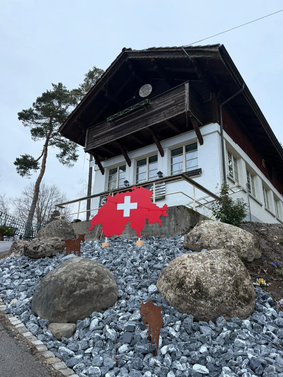 Ein Haus mit Schweizer Flagge und einer Karte der Schweiz im Garten, umgeben von Steinen und Figuren von Kühen.