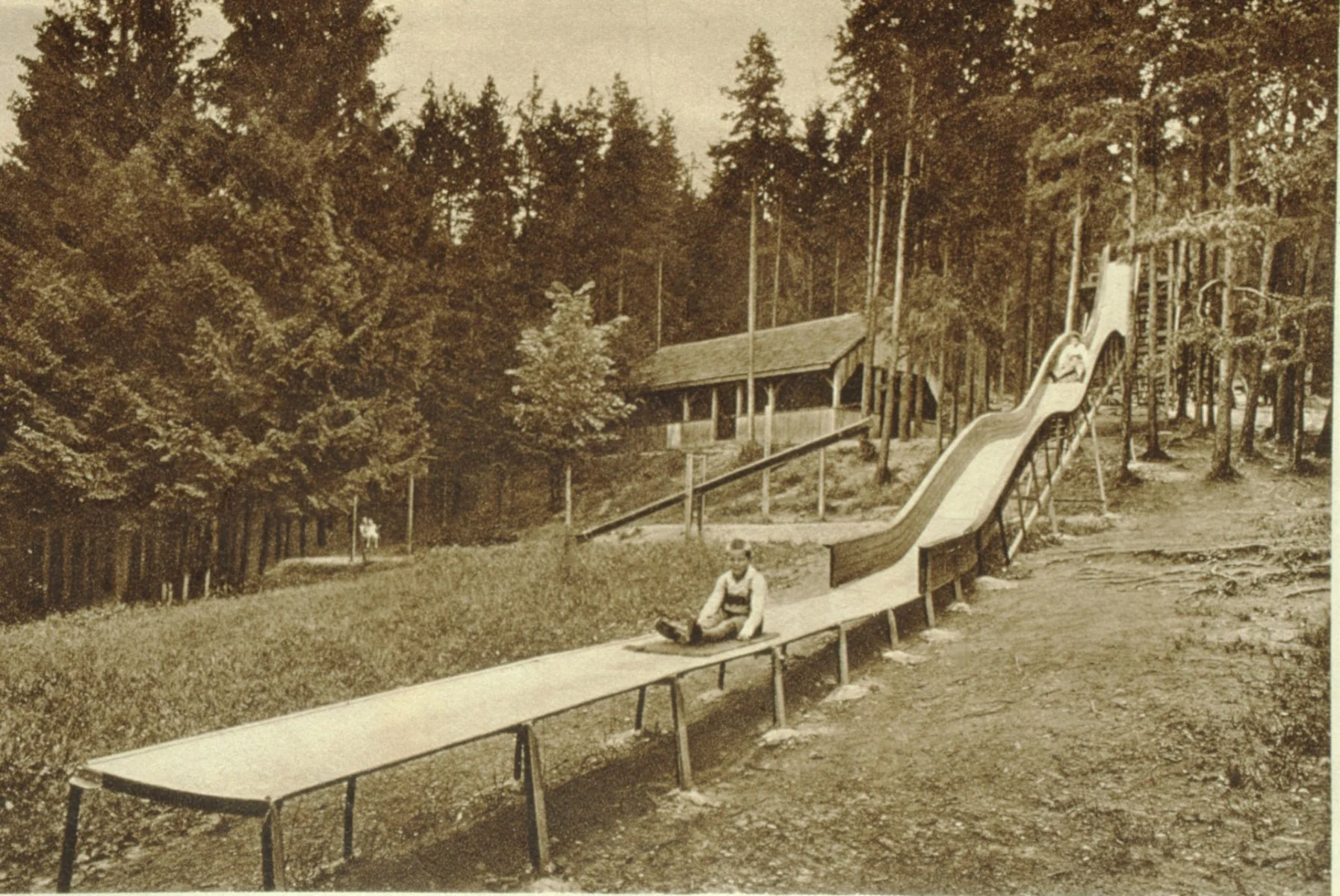 Alte sepiafarbene Fotografie eines Spielplatzes mit einer Rutsche, auf der ein Kind sitzt, umgeben von Bäumen und einem Holzgebäude im Hintergrund.