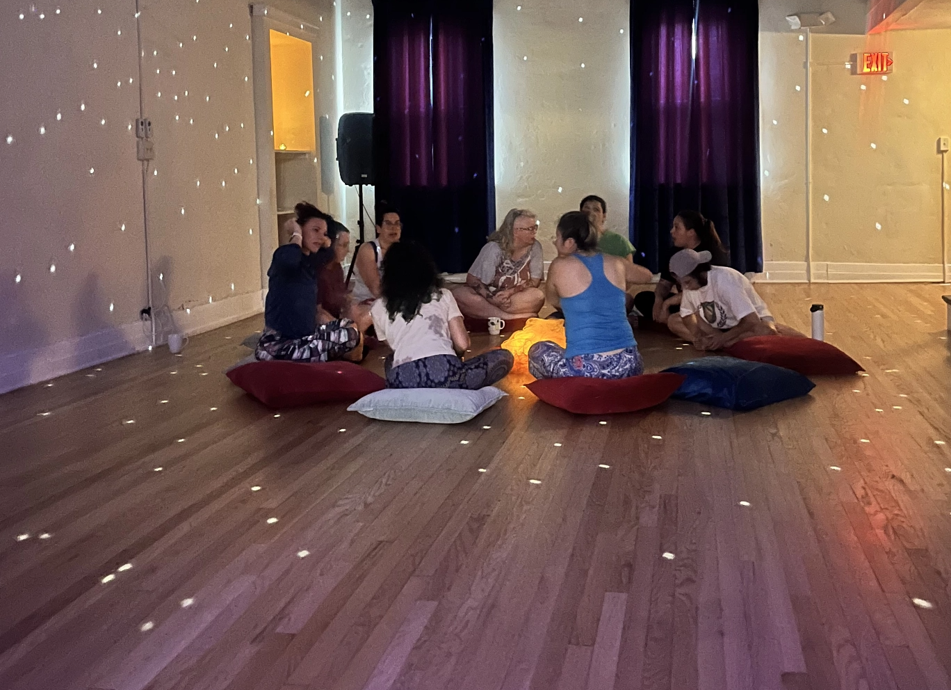 A group of people sitting in a circle on pillows on the floor around a fake fire in the middle.  The lighting is dark and a spotlight is on the disco ball reflecting little specks of light all over the room