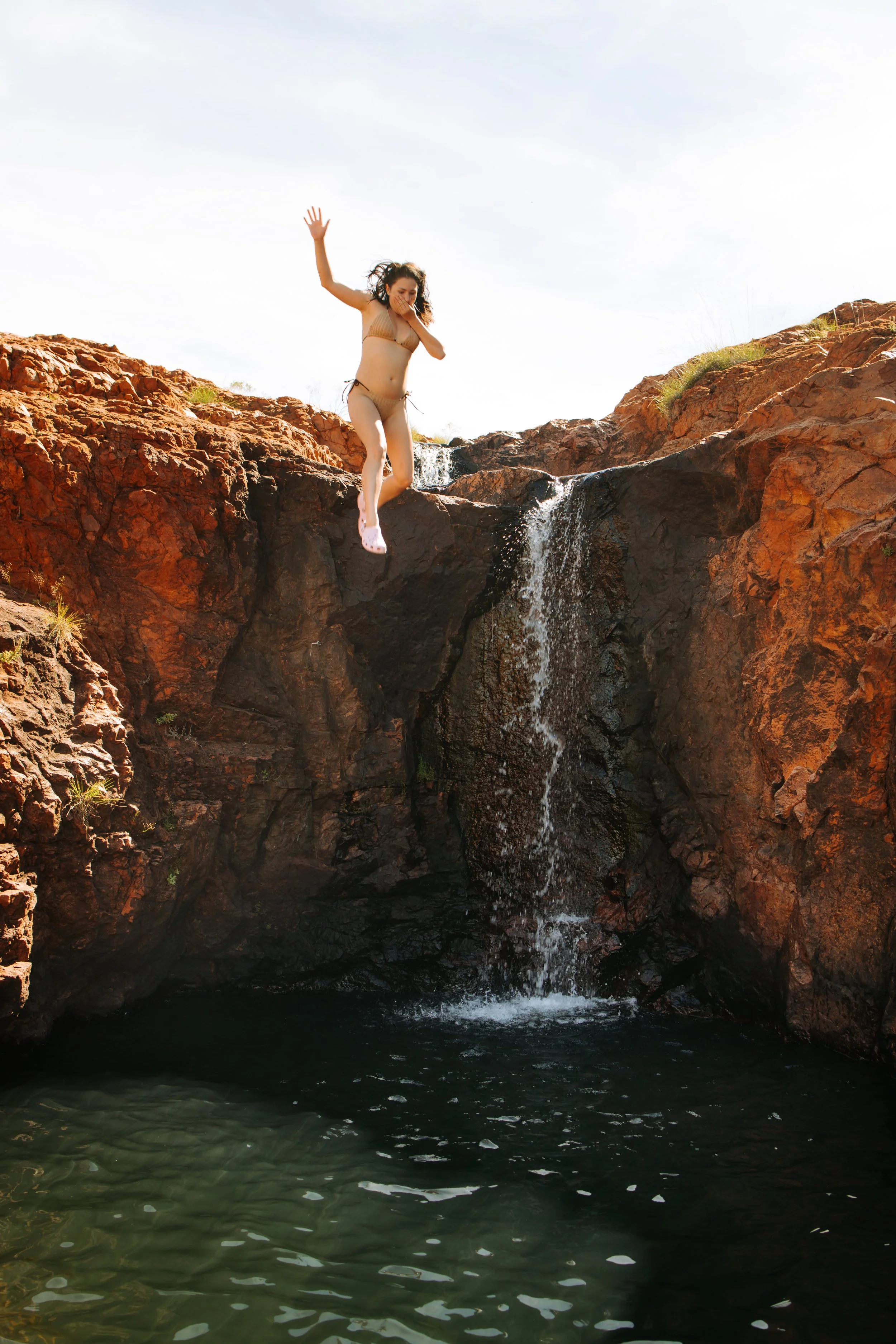 A woman in a beige bikini jumping into a small waterfall and pool in a rocky outdoor setting.