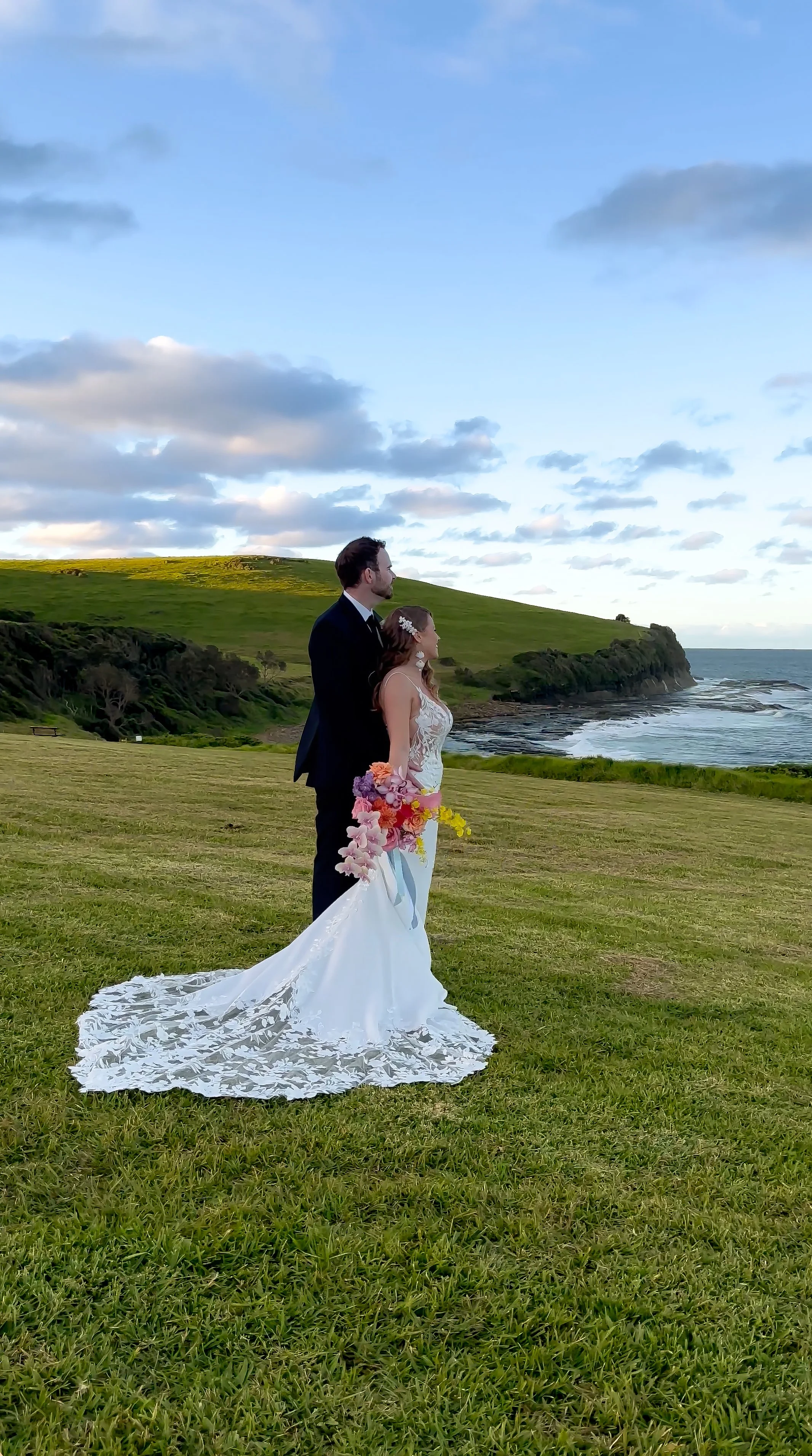 A bride and groom standing on a grassy field by the ocean, with hills in the background under partly cloudy sky.