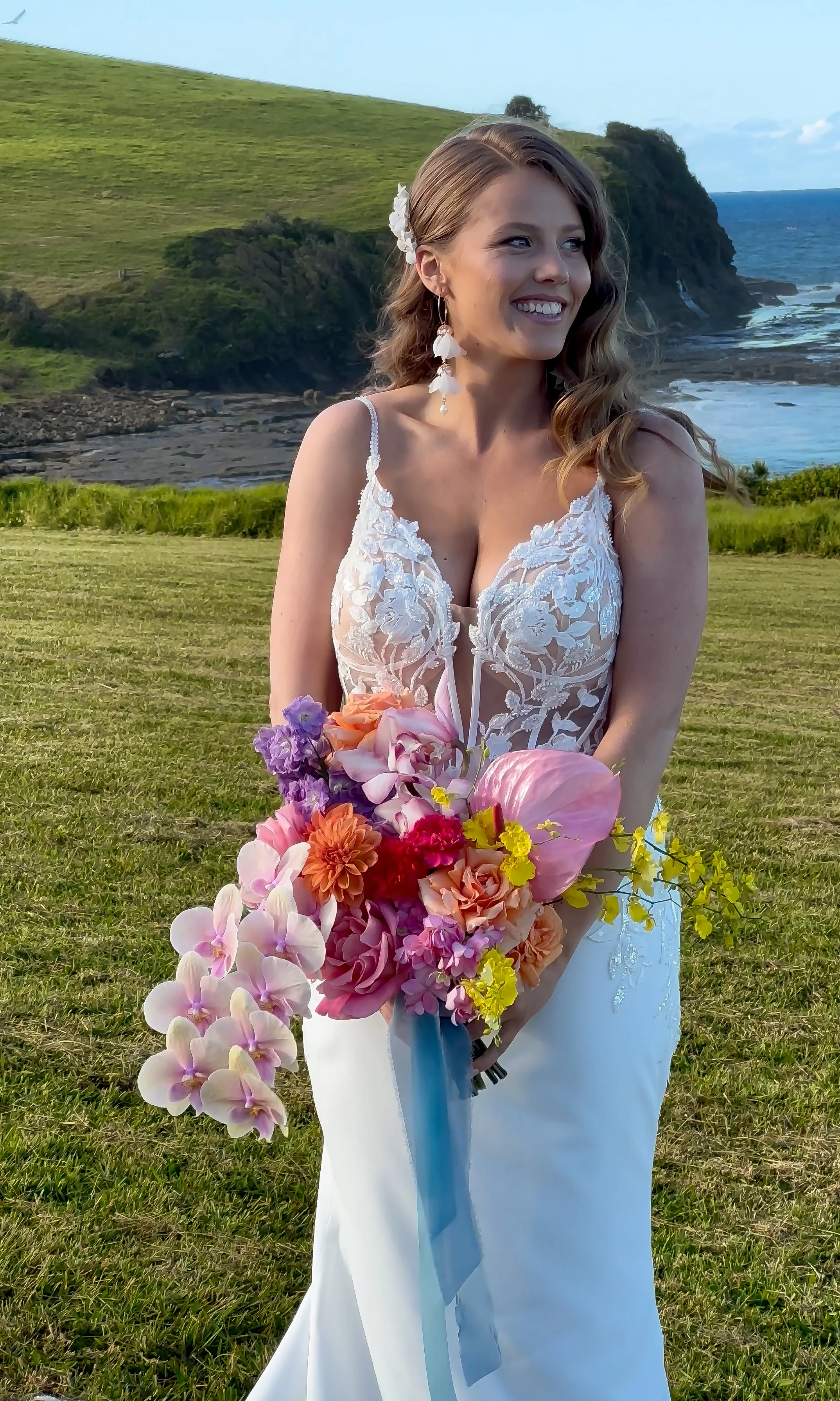 A woman in a white lace wedding gown holding a large colorful bouquet of flowers, standing outdoors near the coast with hills and the ocean in the background.