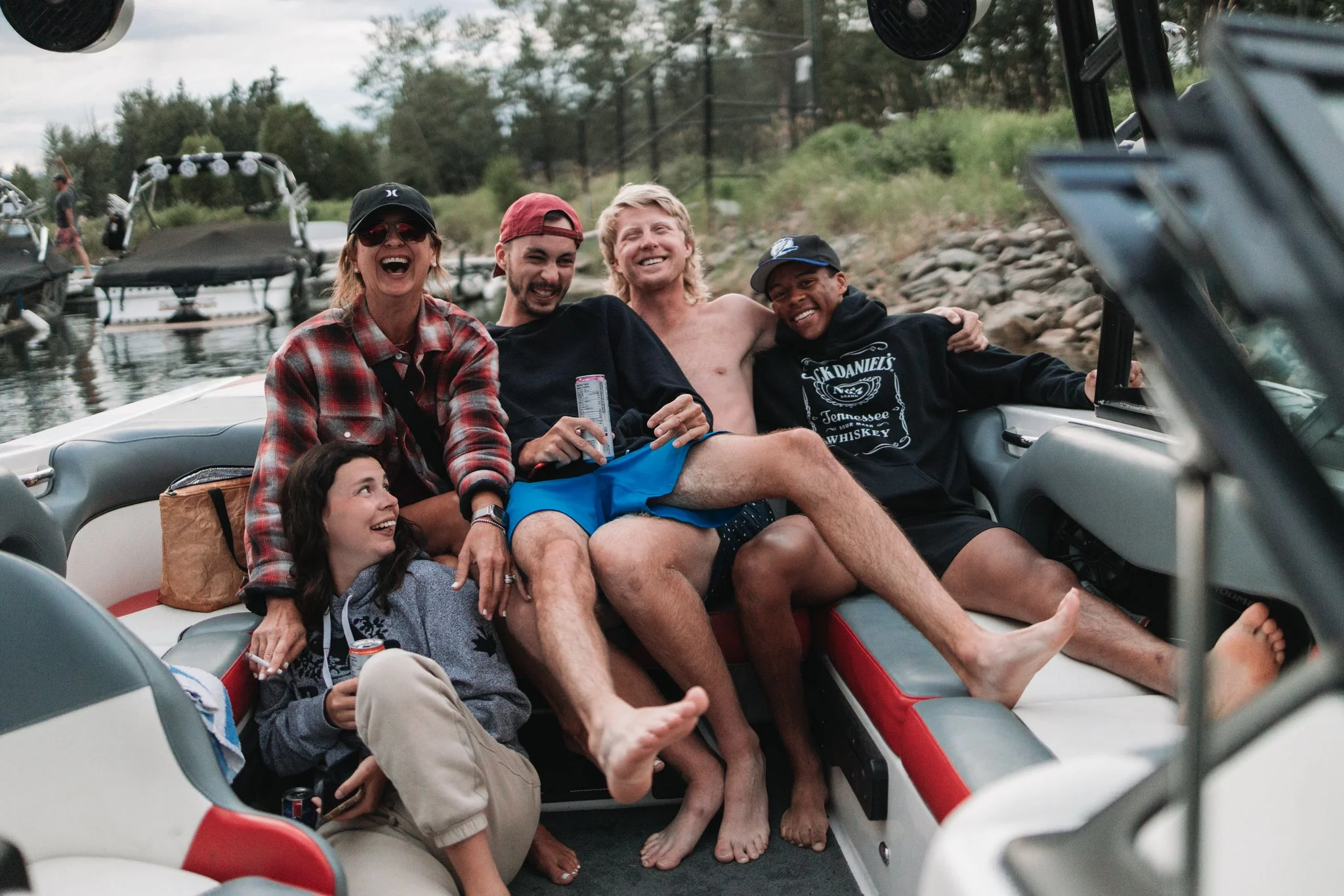 A group of six people sitting on a boat, smiling and enjoying a sunny day. The group includes three women and three men, with some wearing hats and sunglasses. They are near a rocky shoreline with trees, and there are boats and water in the background.