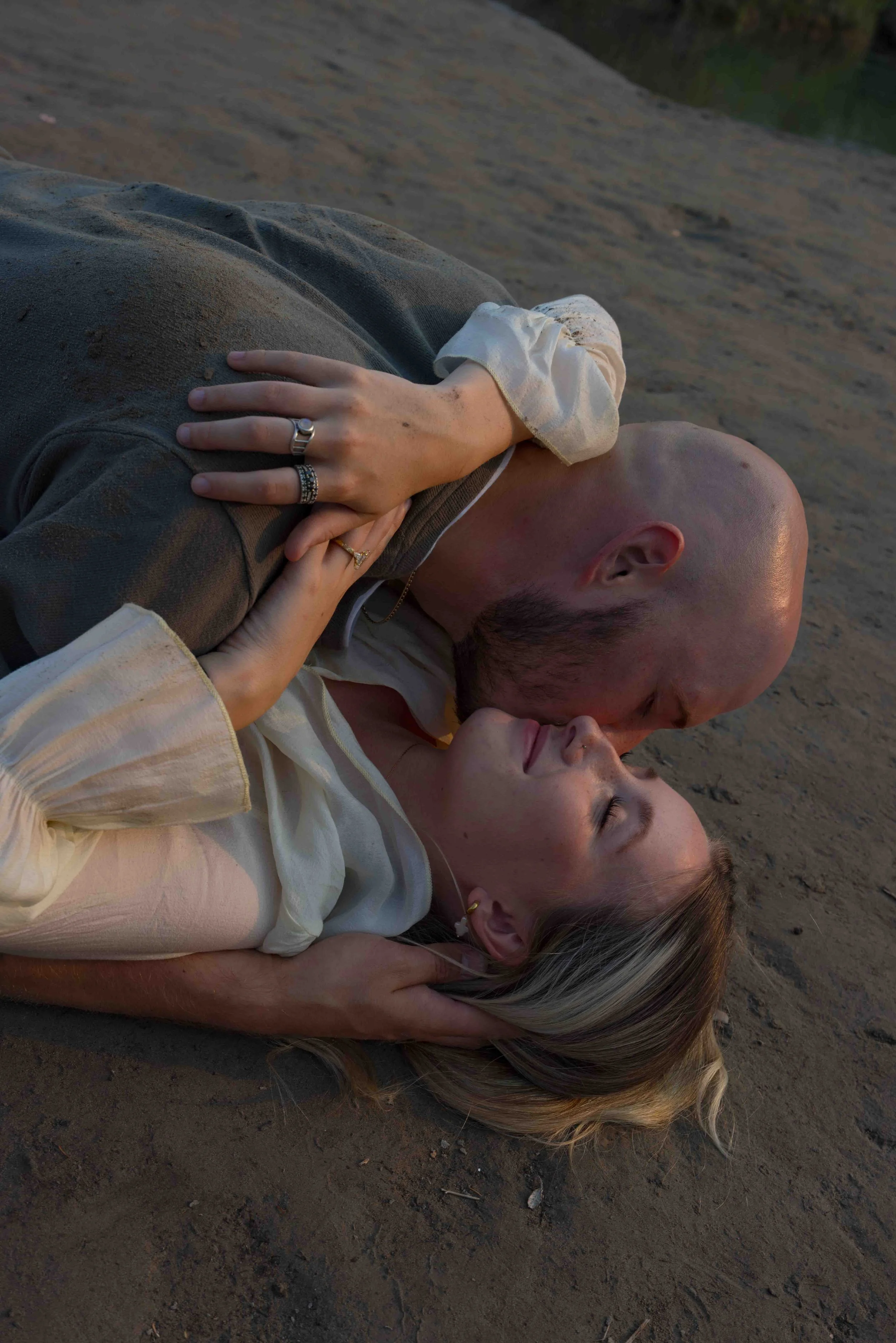 A couple lying on the ground, the woman on top, with sandy soil underneath and a blurred natural background.