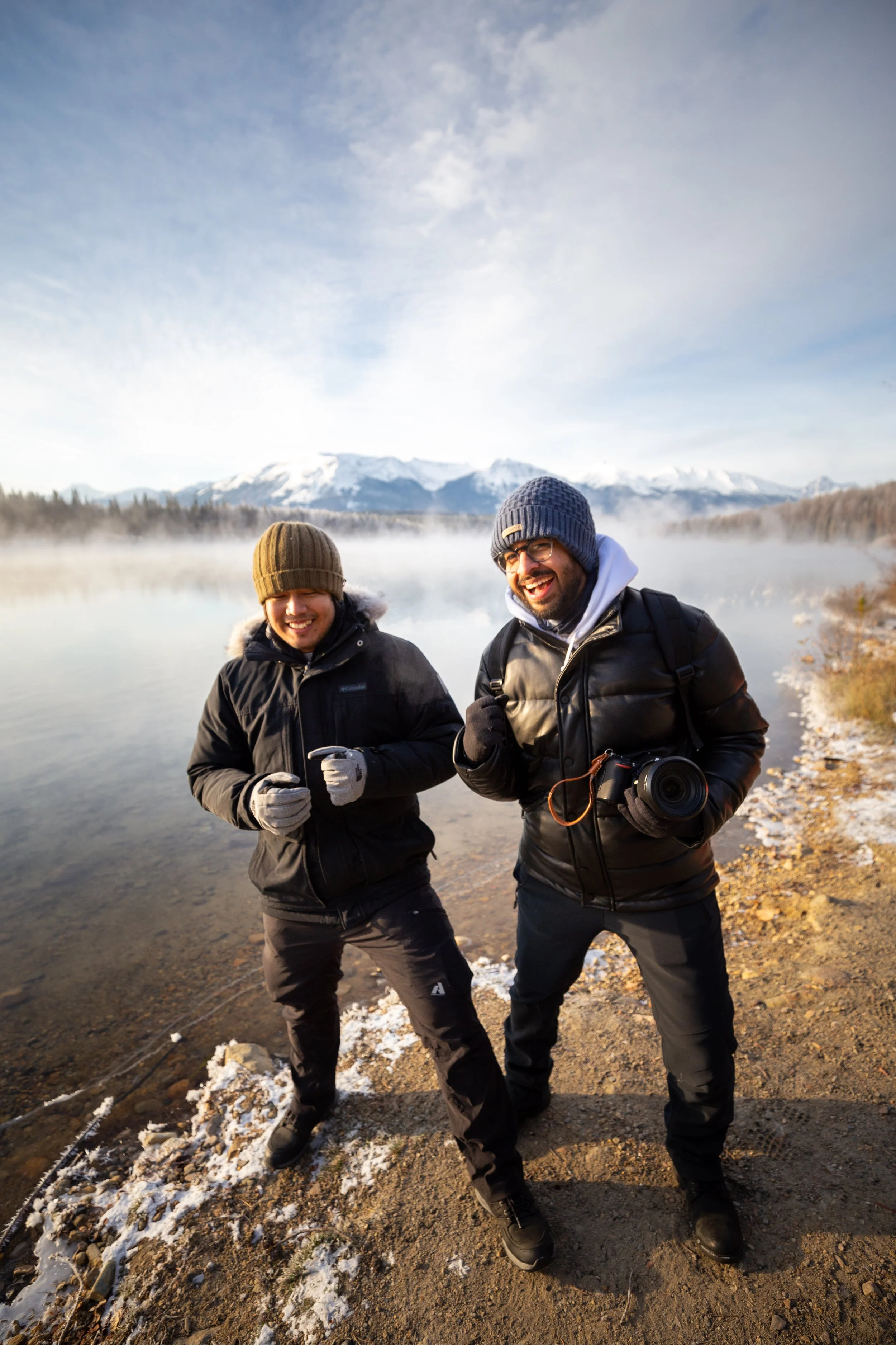 Two smiling men wearing winter jackets and hats, standing near a lake with steam rising from the water, snow-capped mountains in the background, and a partly cloudy sky.