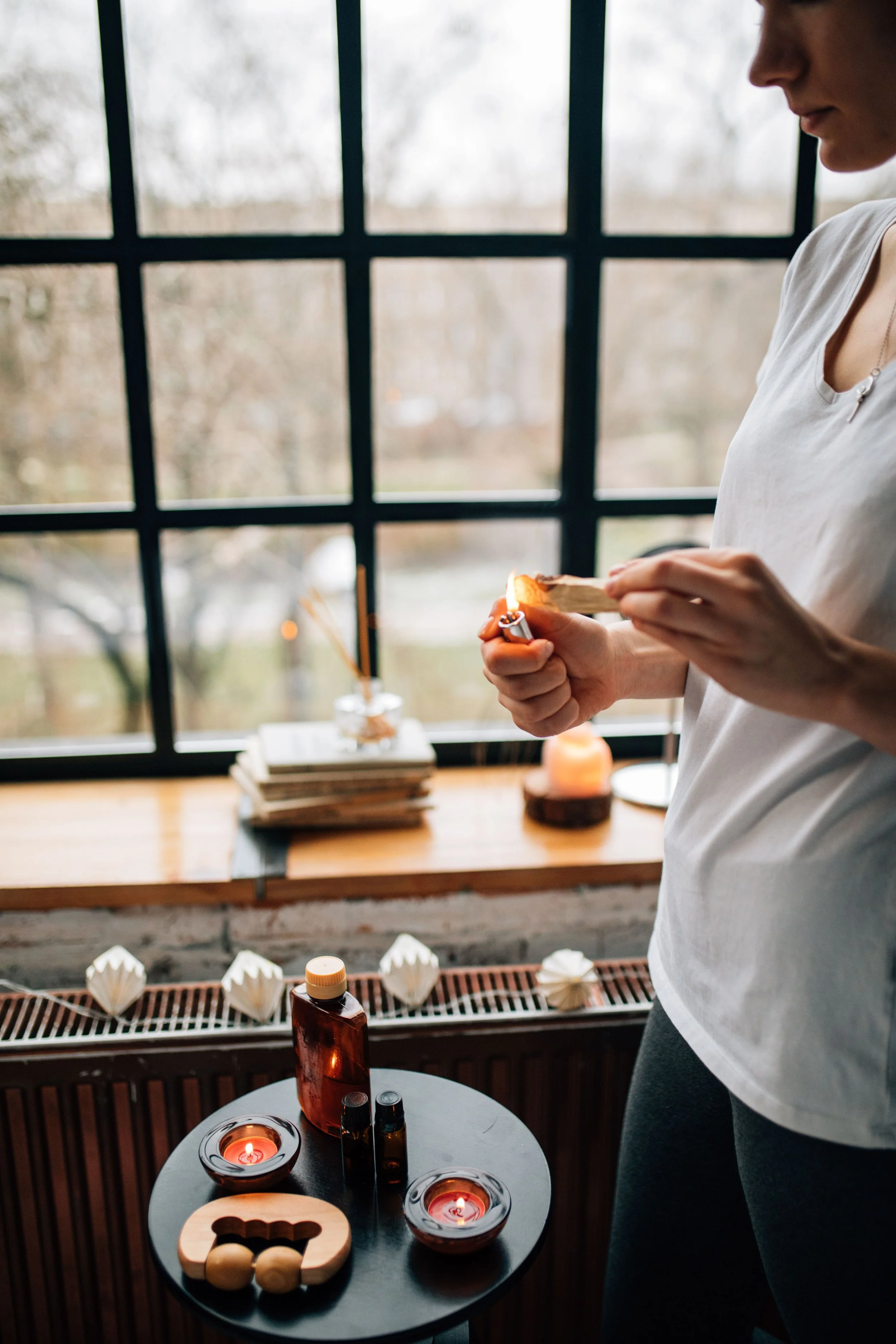 A woman standing near a window with a wooden table in front of her, holding a small object. The table has various items including candles, essential oils, and decorative objects, with a background of a blurred outdoor scene.