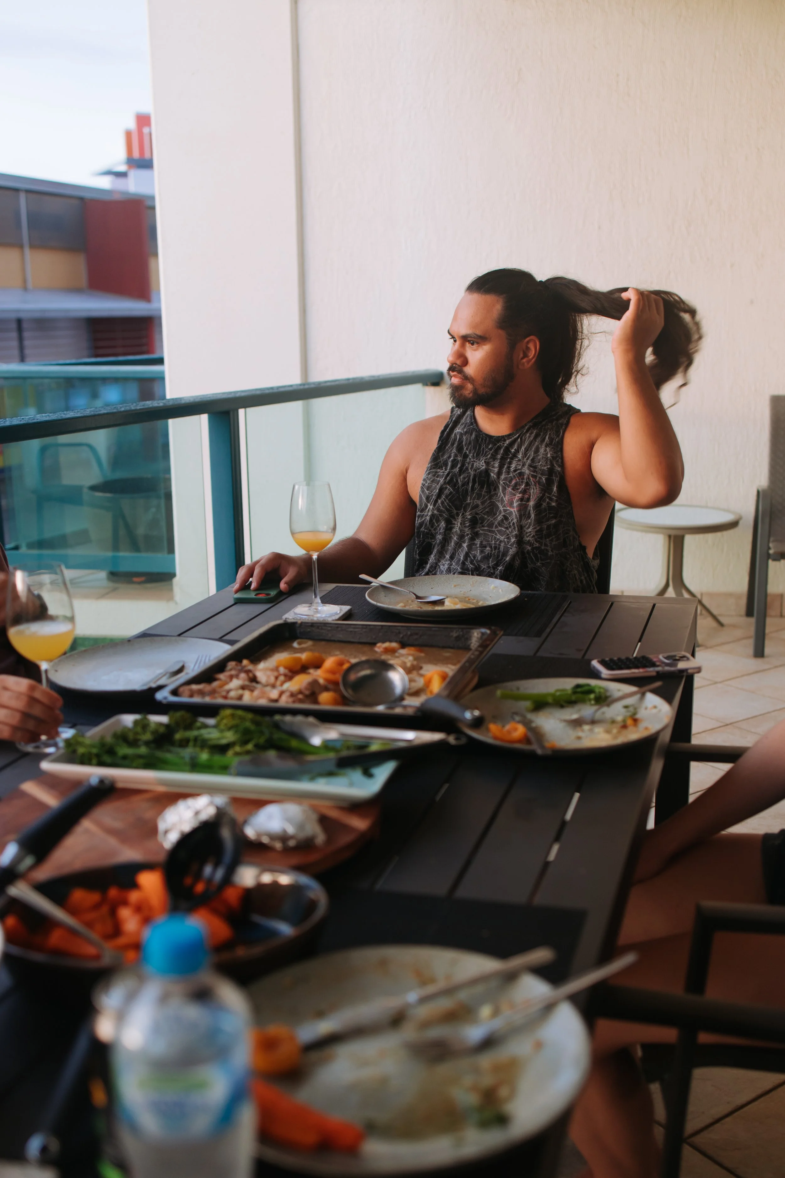 A man with a beard and long hair tied back, wearing a sleeveless shirt, sitting at a dinner table on a balcony, holding his hair with one hand. The table has various plates of food, drinks, and utensils. There are buildings and a cloudy sky in the background.
