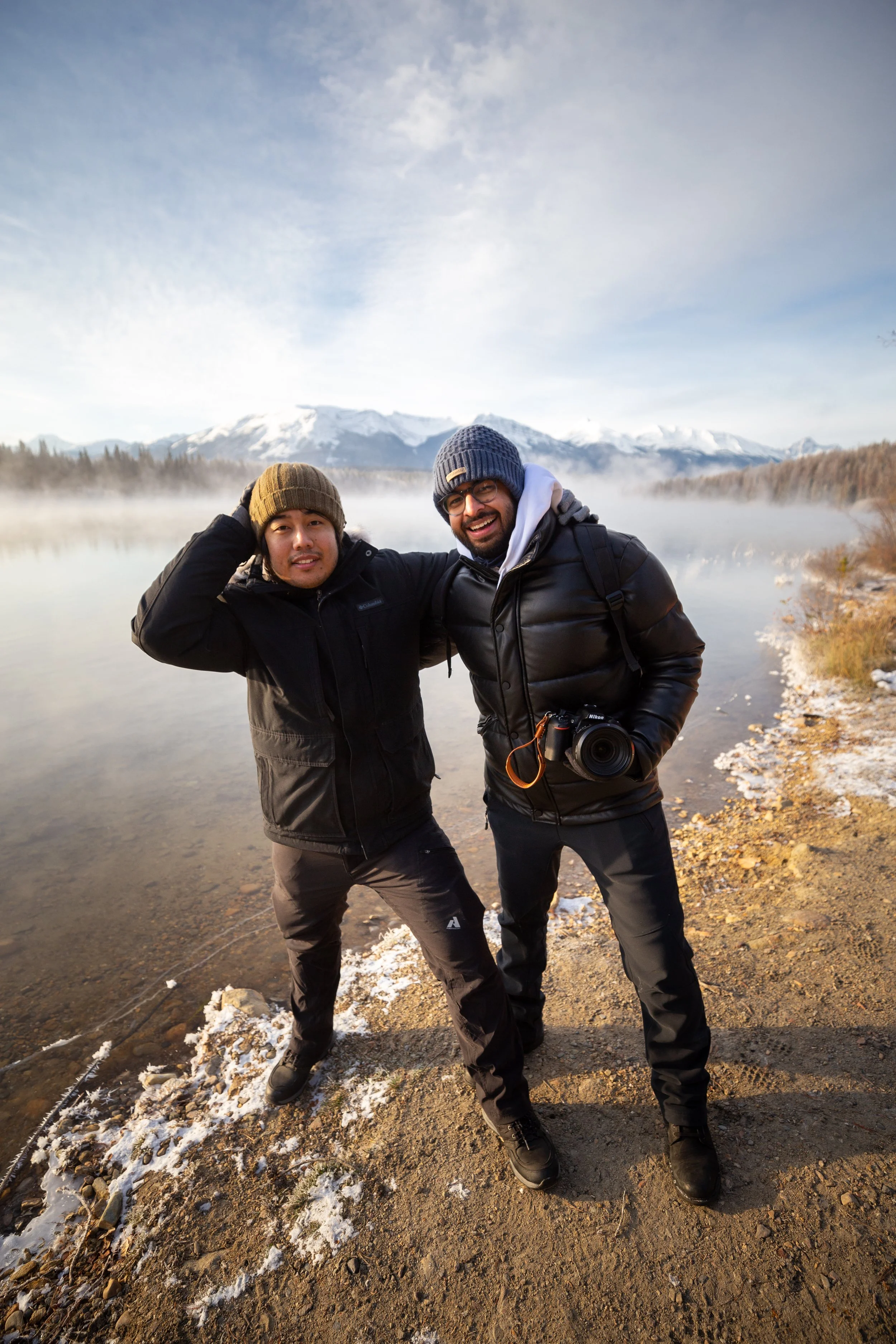 Two men standing on a rocky and snowy shoreline near a body of water, with snow-capped mountains and a partly cloudy sky in the background. One man has his hand on his head, and the other holds a camera.