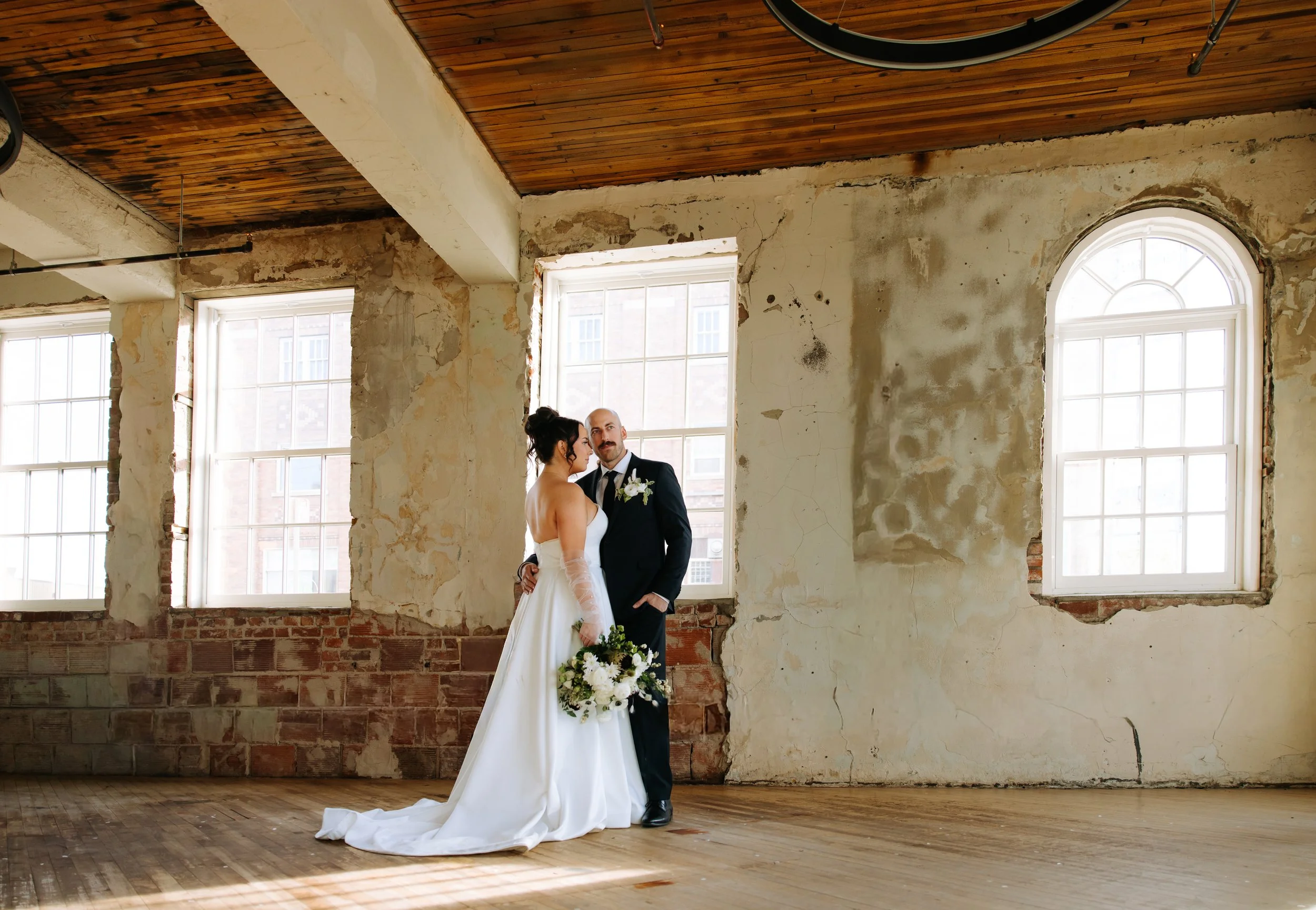Bride and groom photographed inside a bright industrial wedding venue with large windows and textured walls.