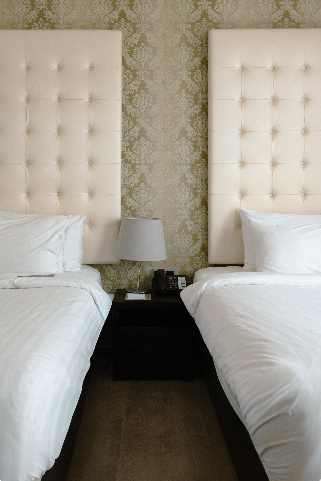 Two hotel beds with white bedding and large tufted cream-colored headboards separated by a nightstand with a lamp, telephone, and notepad, against patterned wallpaper.