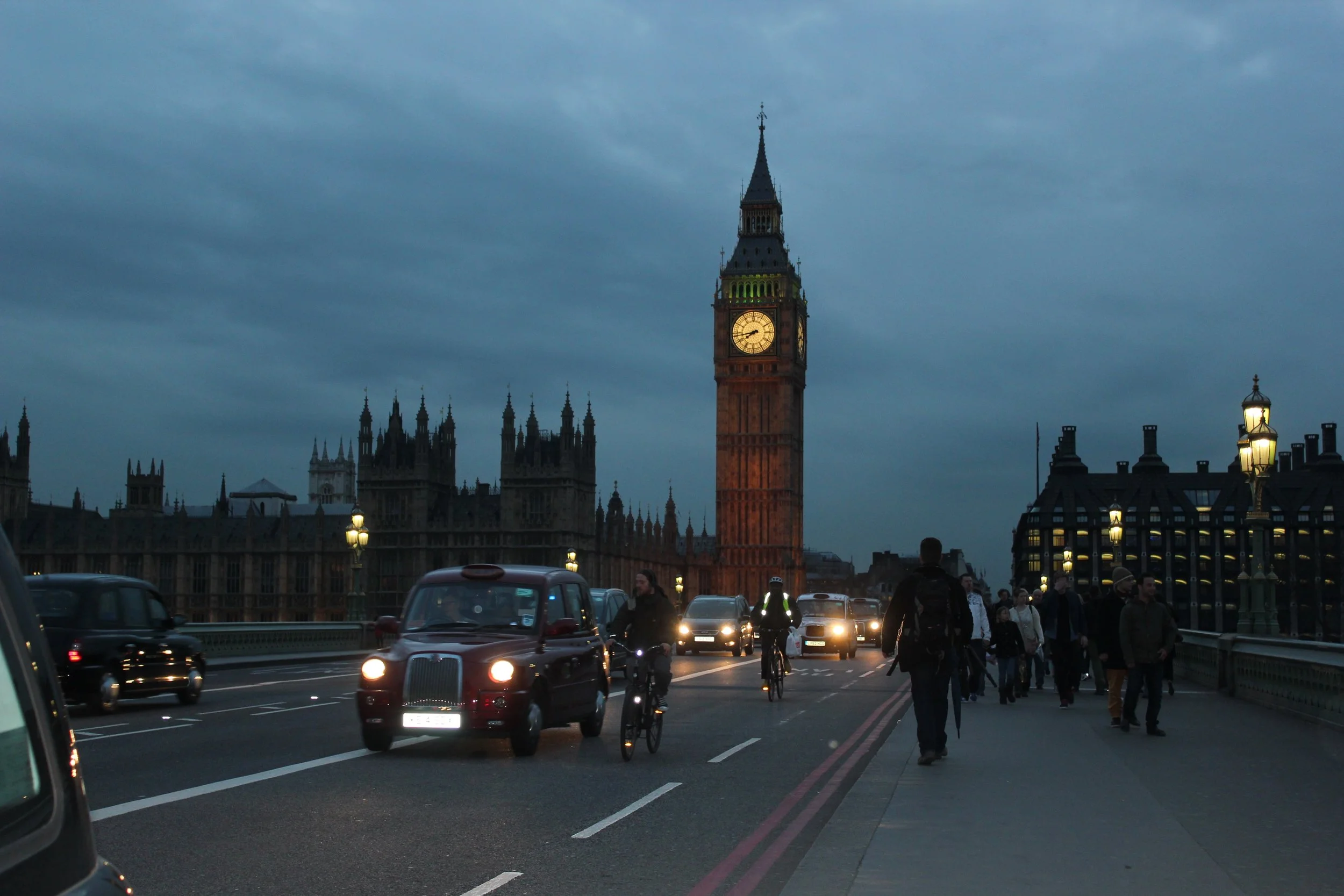 View of Big Ben clock tower in London at dusk with people walking, cycling, and cars on the bridge.