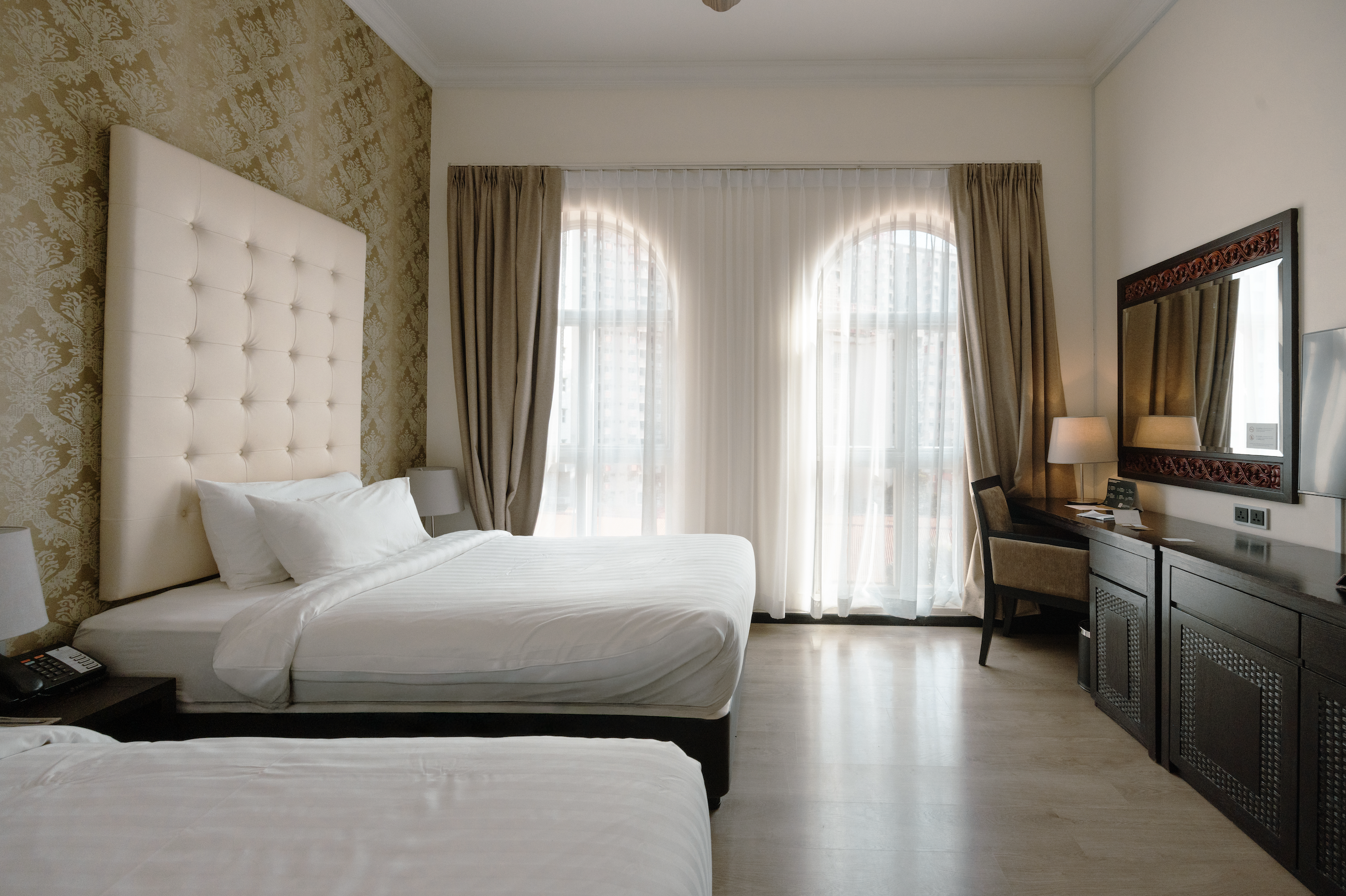 A hotel room with a large bed, a bedside table with a remote, a window with sheer white and beige curtains, a mirror, and a desk with two lamps and a chair.