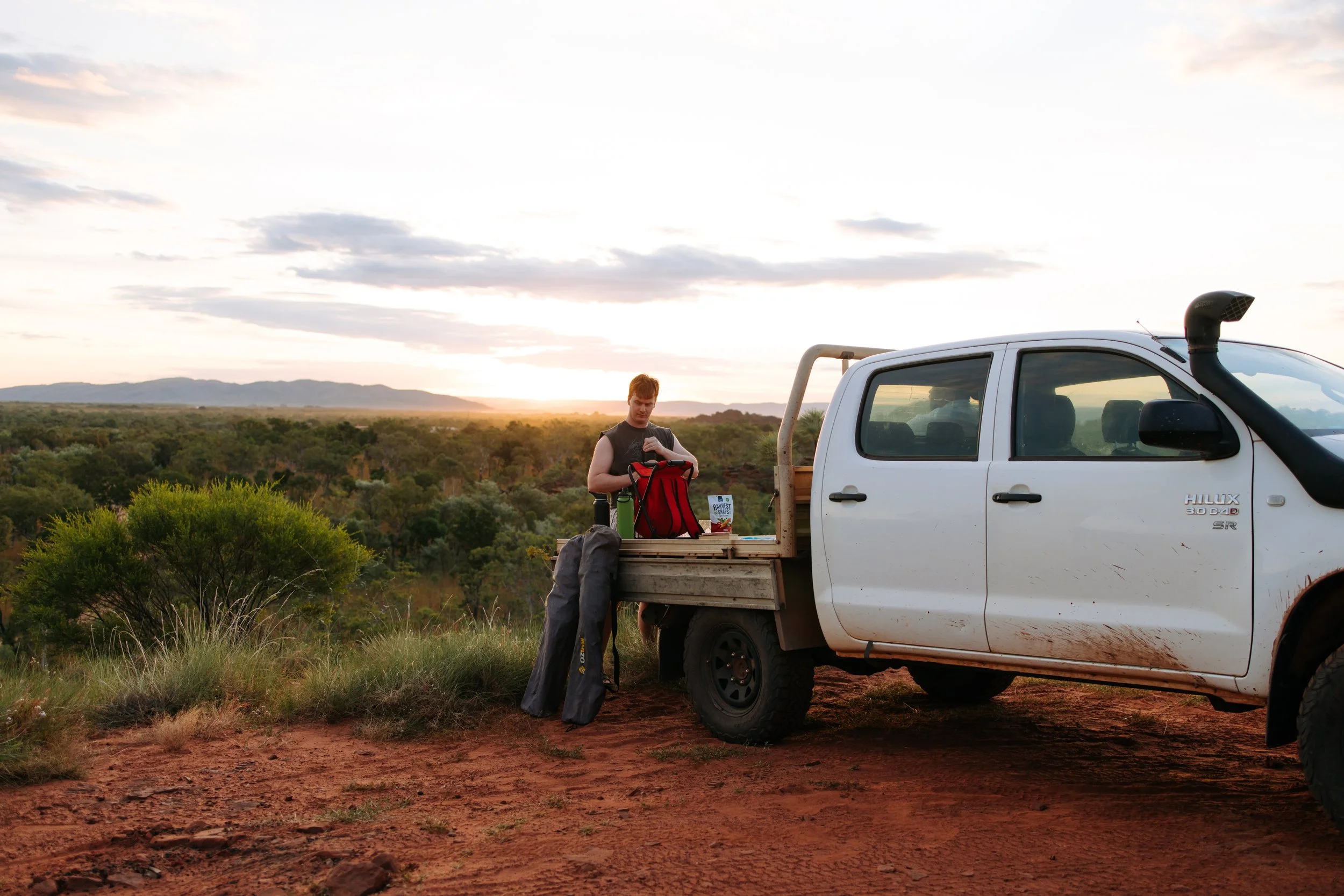 A person standing next to the open back of a white pickup truck in a rural landscape during sunset. The person is packing or unpacking a red backpack, with a green water bottle and other gear nearby. The background features green bushes and distant mountains under a partly cloudy sky.