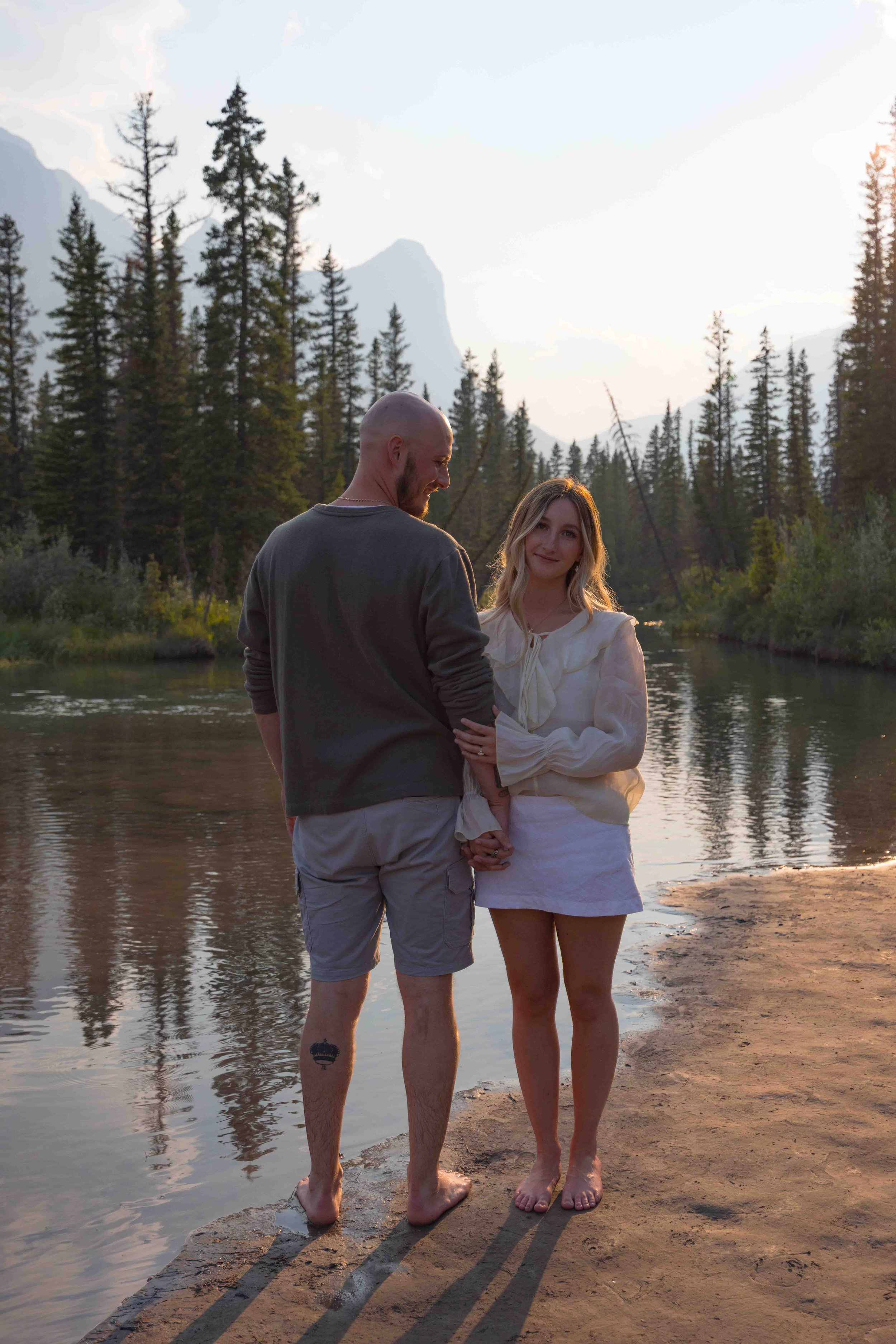 A couple standing at the edge of a river in a forested mountain landscape during sunset. The man is bald with a beard, wearing a dark long-sleeve shirt and shorts, with a tattoo on his leg. The woman has long blonde hair, wearing a white blouse and s