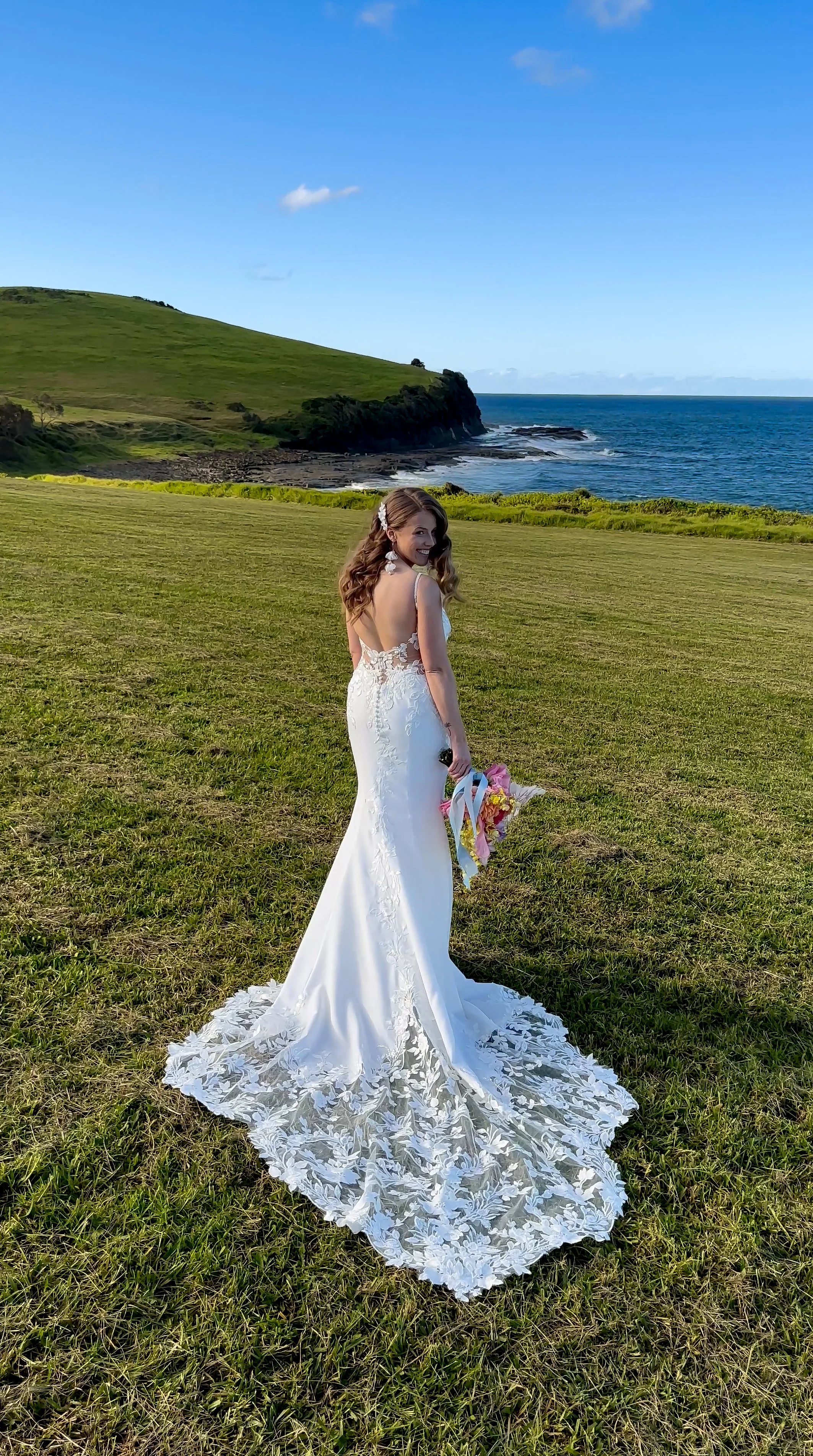 A woman in a white wedding dress holding a bouquet of flowers, standing on a grassy field near a coastline with green hills and blue ocean under a clear sky.