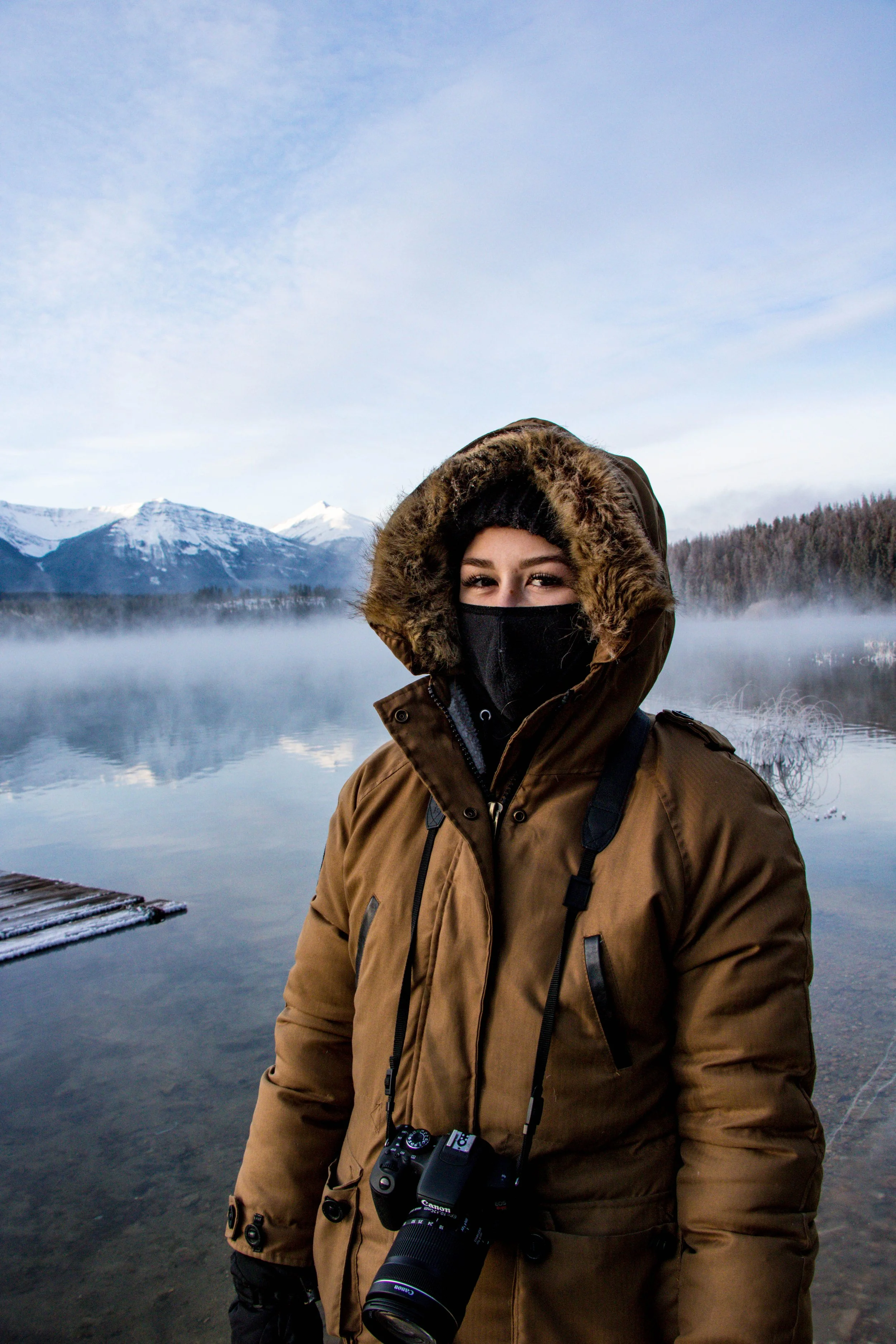 Person wearing a brown winter coat with a fur-lined hood and a black face mask, standing near a frozen lake or river with snow-capped mountains and a cloudy sky in the background.