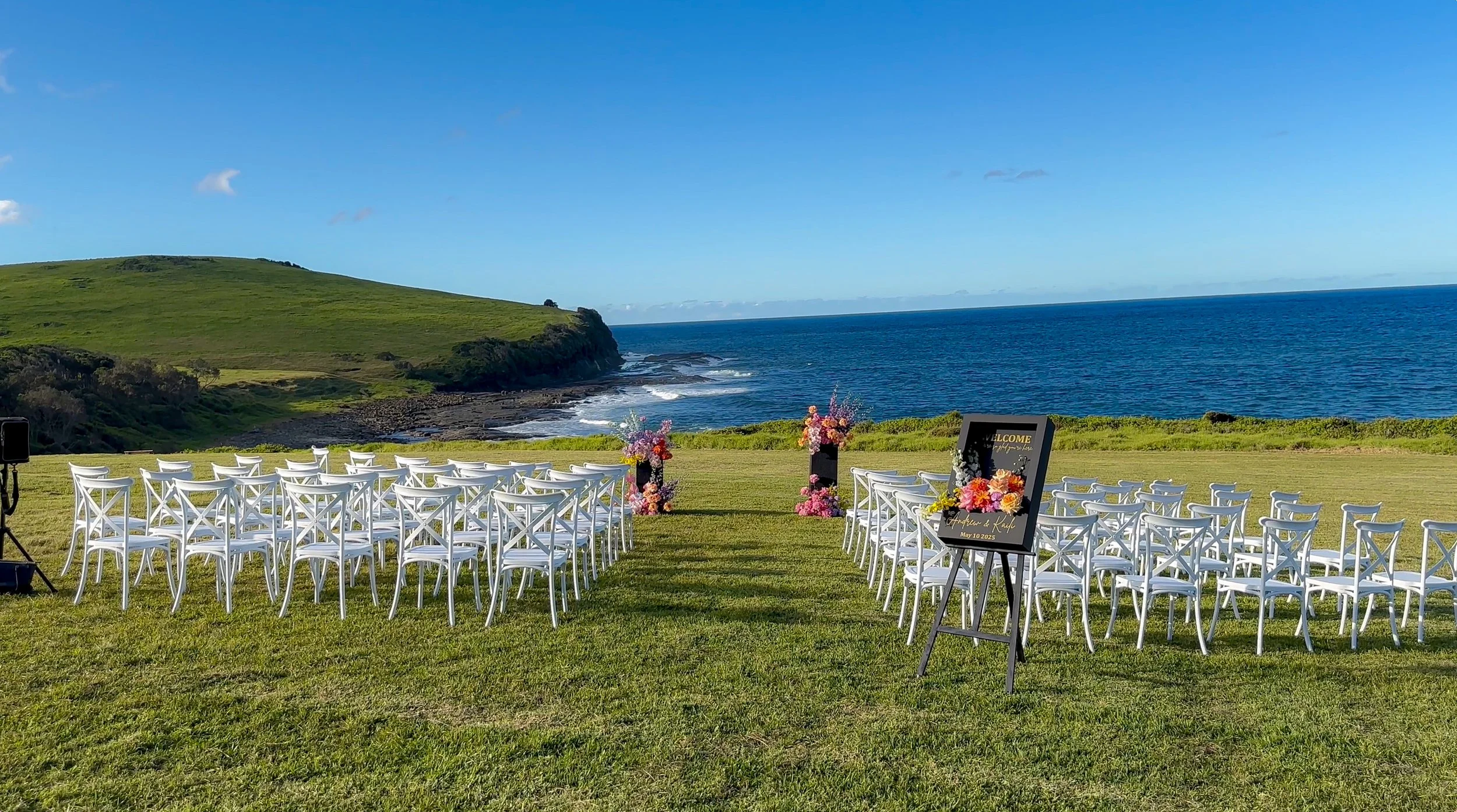 An outdoor wedding ceremony setup on a grassy area overlooking the ocean, with white chairs arranged in rows on either side of a decorated aisle, floral arrangements, and a welcome sign, under a clear blue sky with green hills in the background.