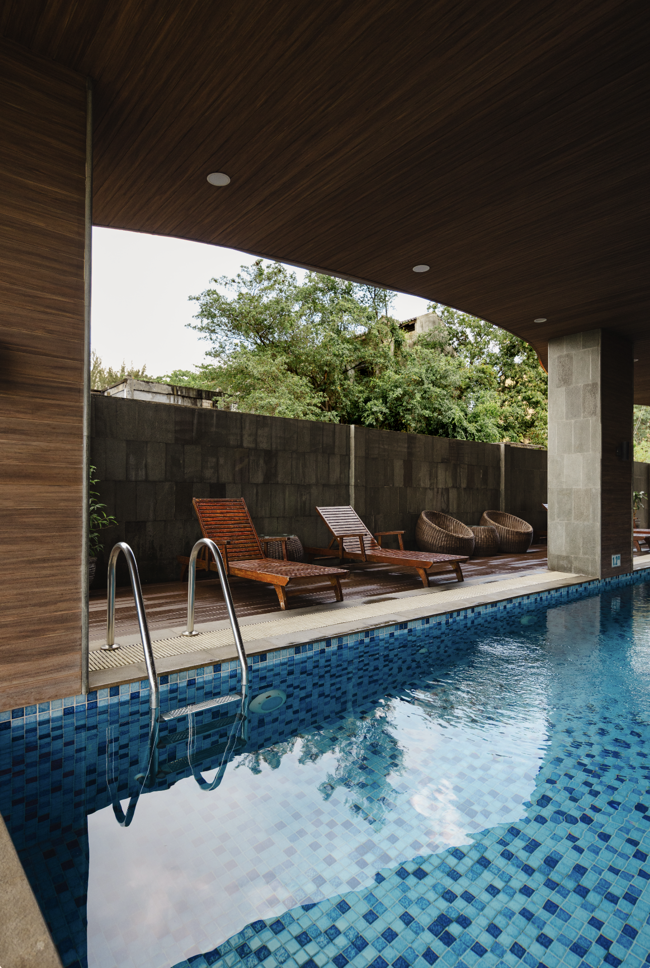 Swimming pool area with wooden lounge chairs, wicker chairs, and a dark privacy wall, covered by a wooden ceiling with built-in lights.