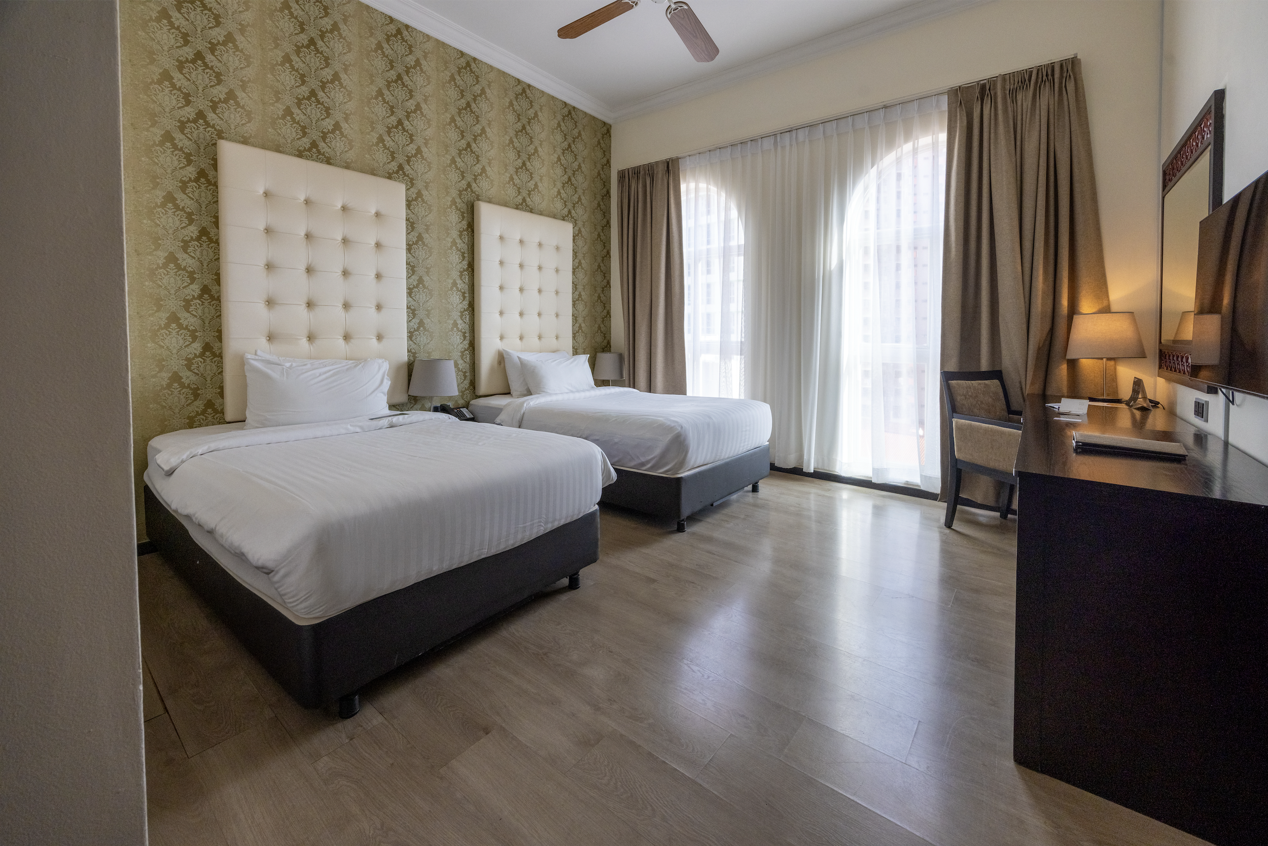 A hotel room with two twin beds with white linens and tufted headboards, a window with sheer and blackout curtains, a desk, a chair, and a wall-mounted TV.