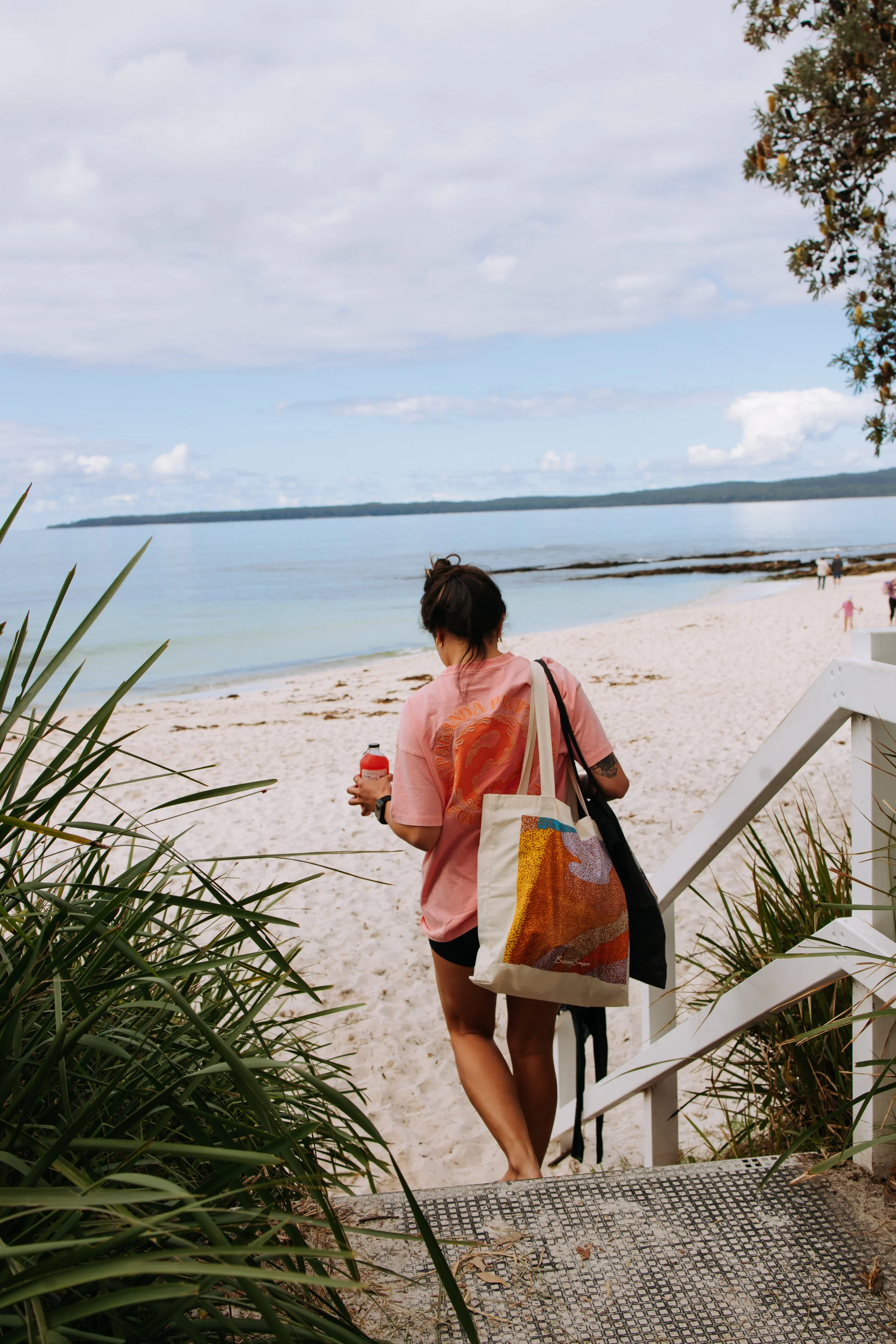 A woman with dark hair tied in a bun, wearing a pink T-shirt and black shorts, is walking down white stairs onto a sandy beach. She is carrying a tote bag with a colorful abstract pattern on her shoulder and holding a drink in her right hand. The scene includes green plants on the sides, a calm body of water, a cloudy sky, and a few people in the distance.