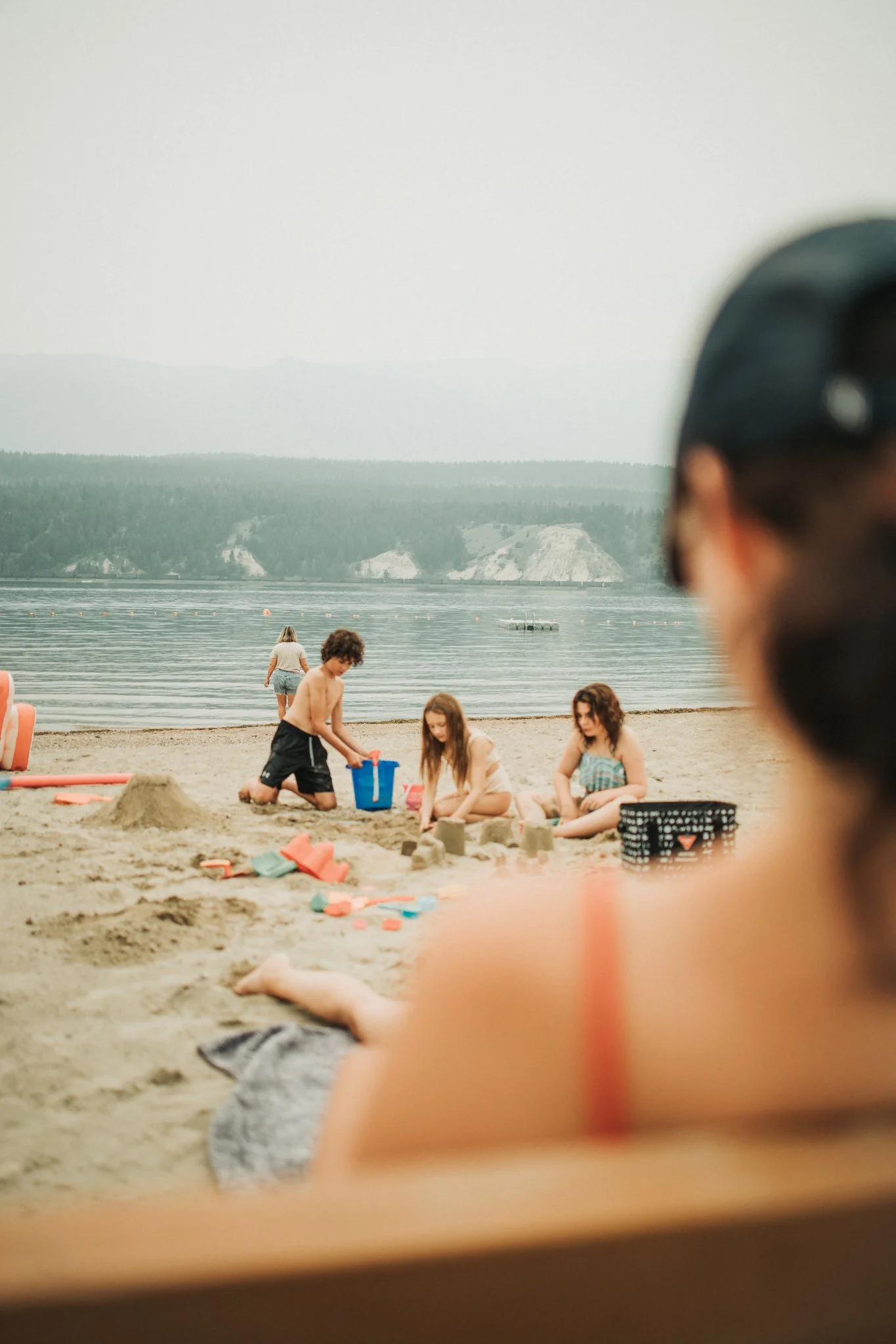 Children playing and building sandcastles on a beach with boats on the water and mountains in the background.