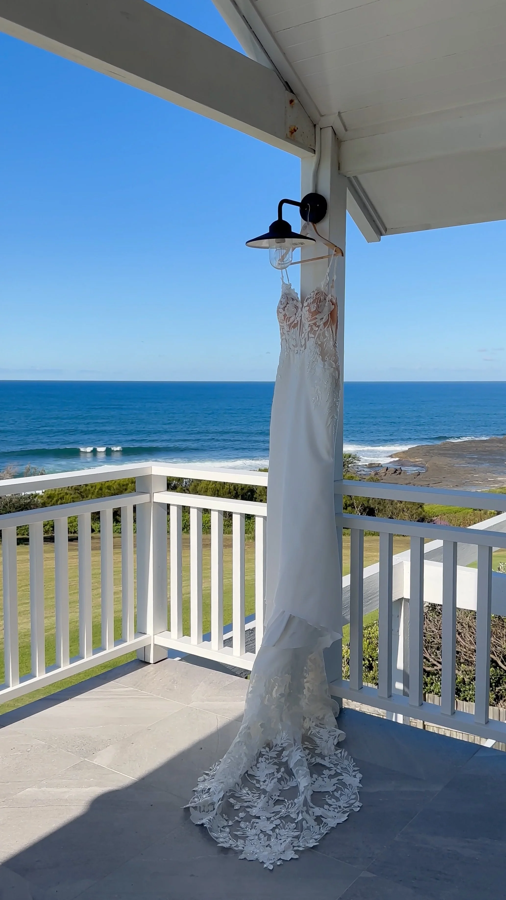 A white wedding dress hanging on a black lamp fixture on a porch with a view of the ocean, blue sky, and green landscape.
